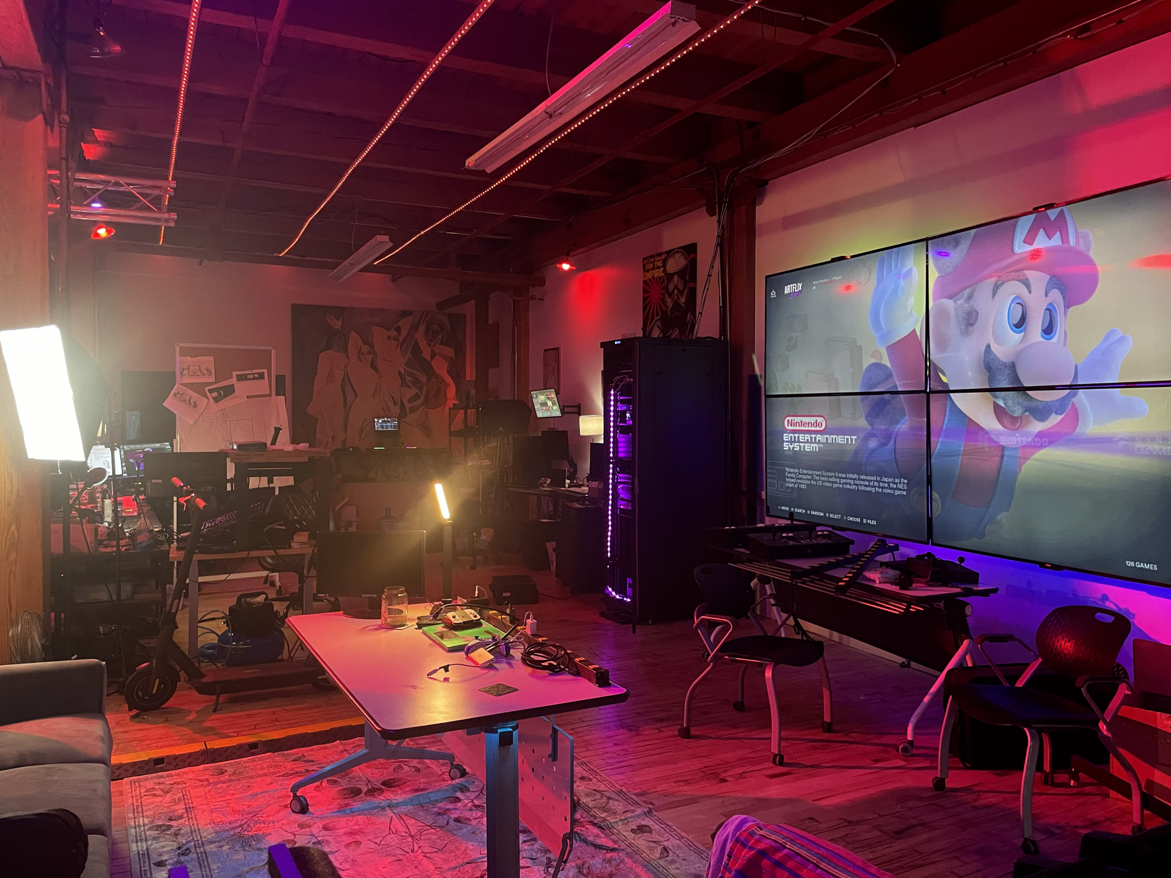 The image shows a dimly lit gaming studio with various equipment, a large screen displaying a Nintendo game, and a table in the center.