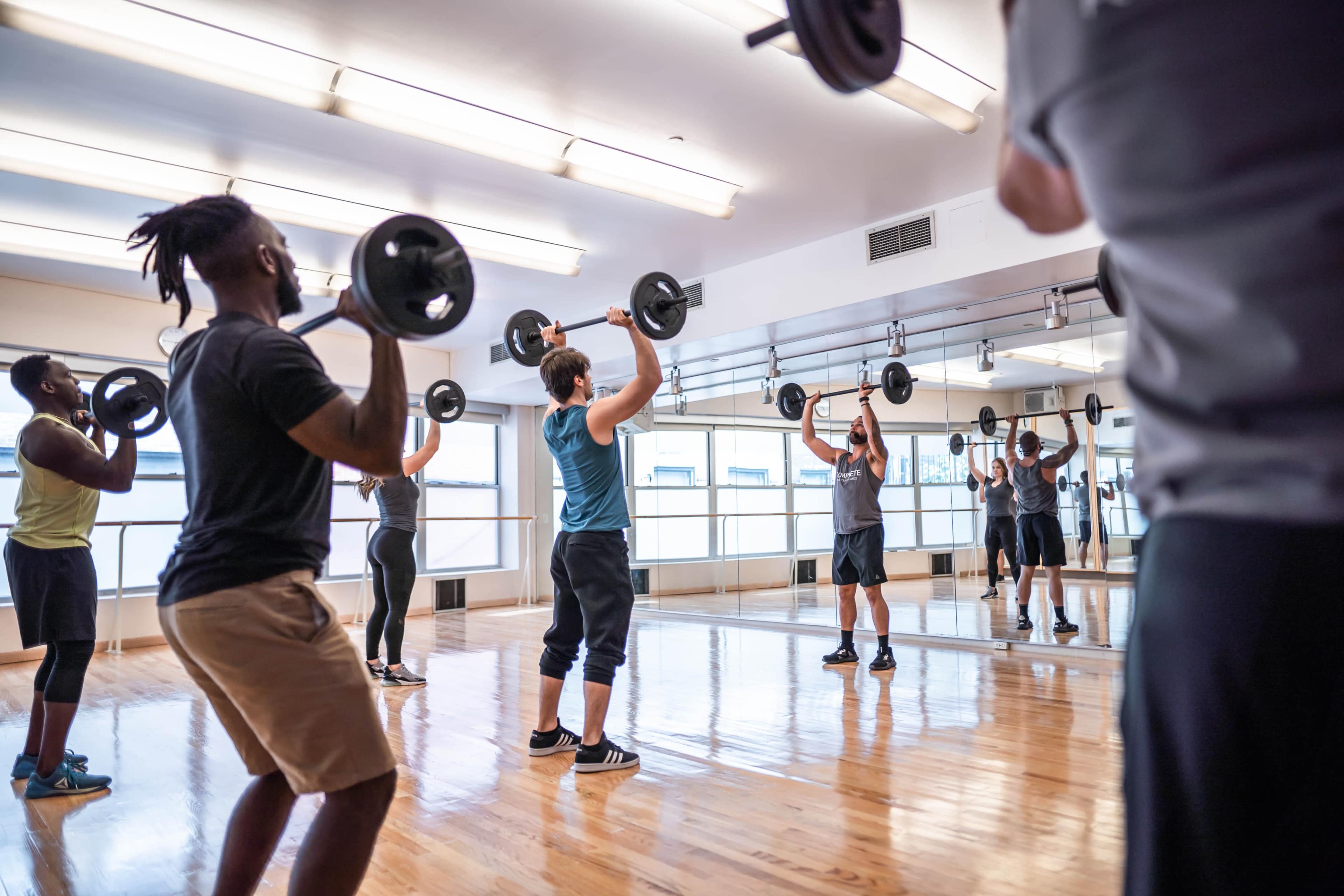 A group of individuals performs weightlifting exercises in a bright gym studio with mirrors.