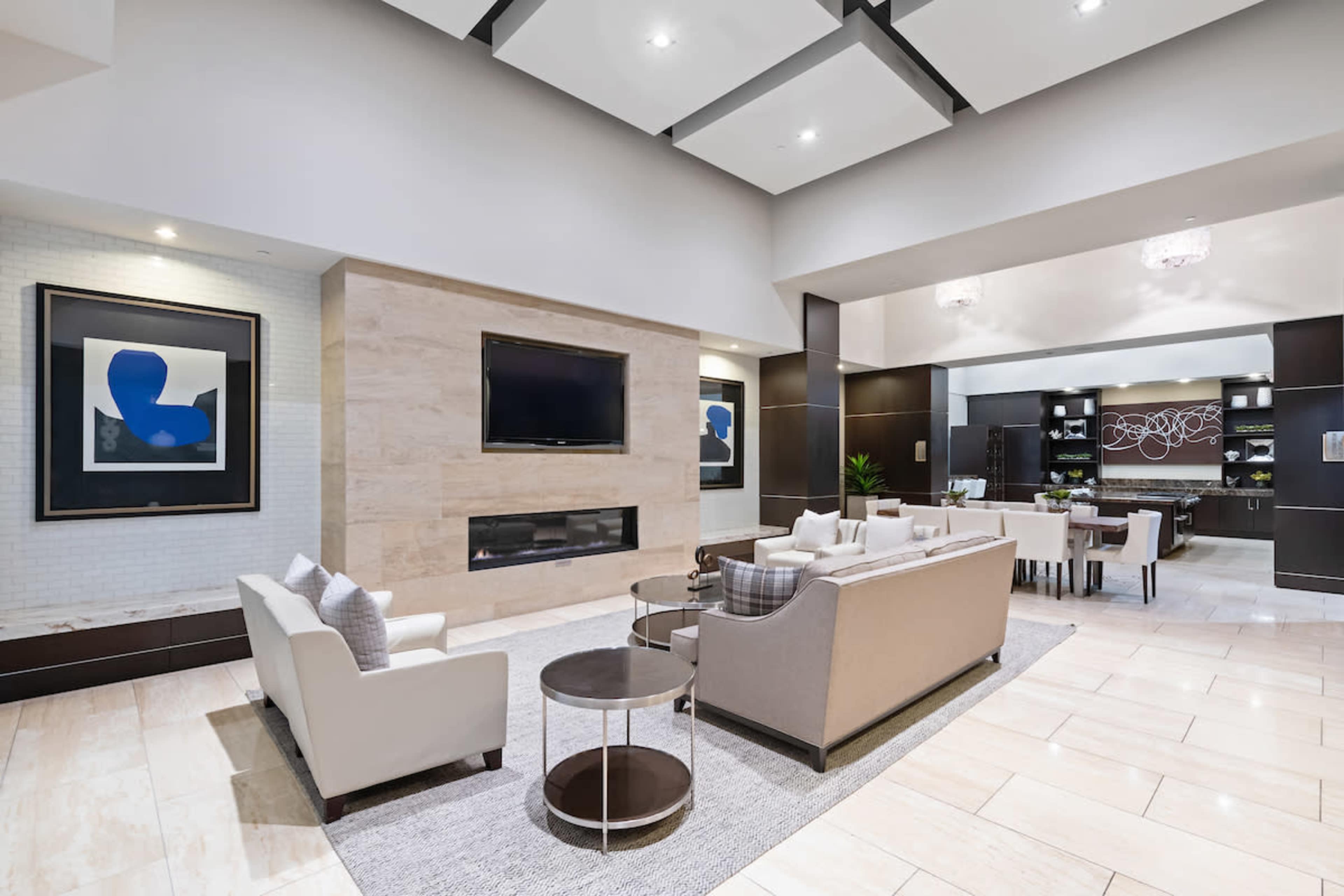 A modern lounge area with a light-colored sofa set, a circular coffee table, a large television mounted on a stone wall, and a dining area in the background.