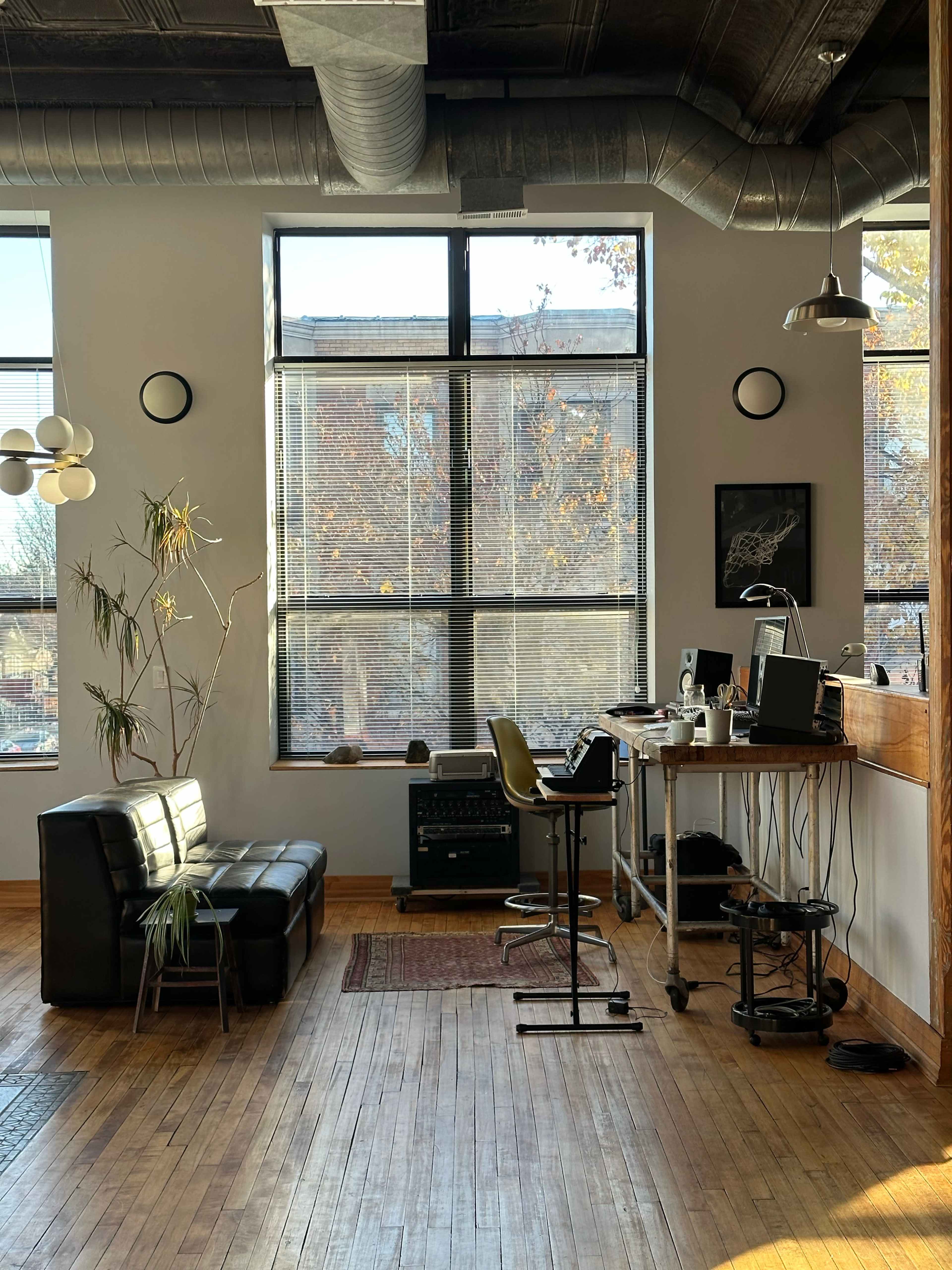 The image shows a modern office space with a black sofa, a desk with a computer, and large windows allowing natural light to enter.