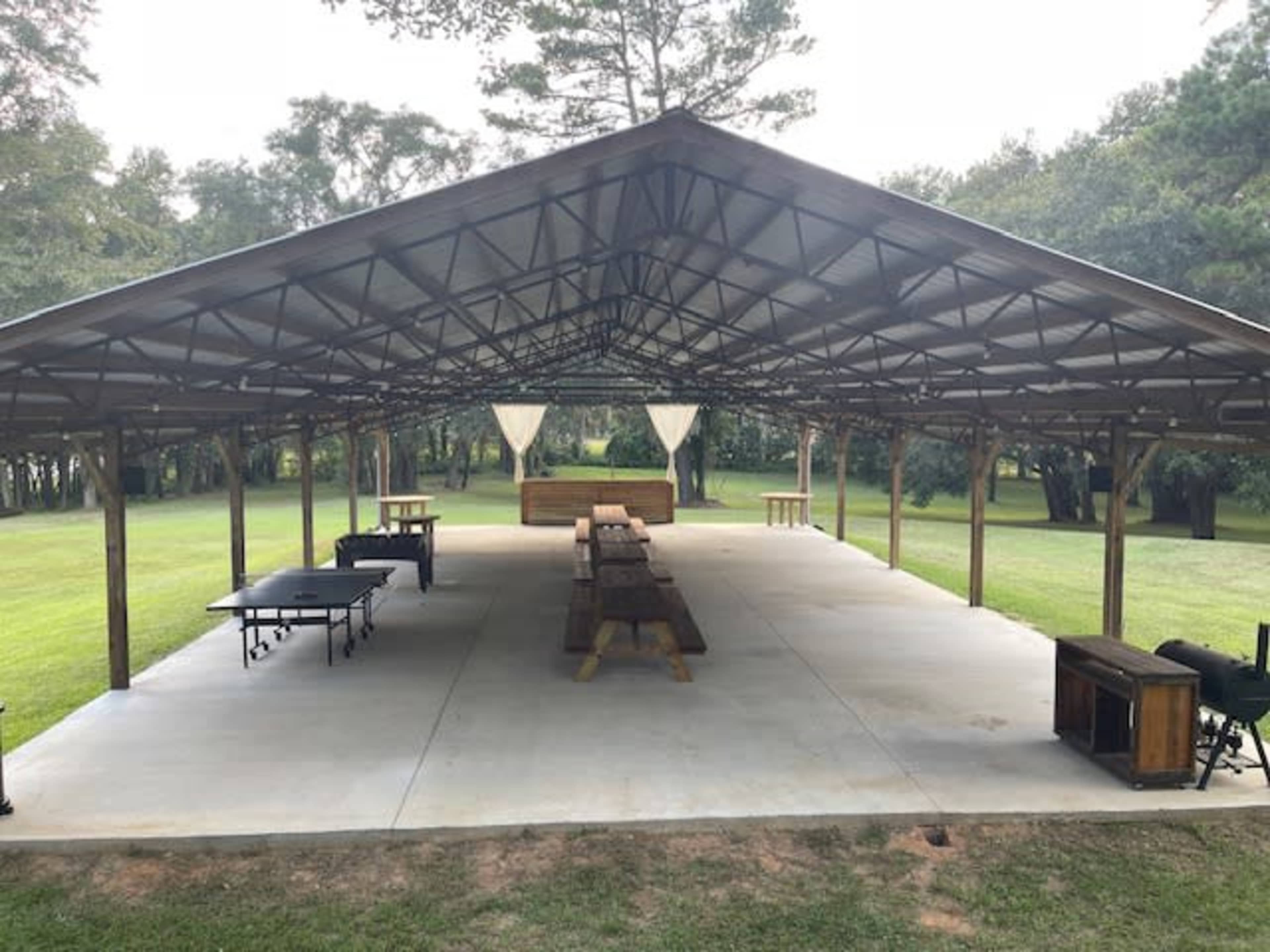 A large open pavilion with a concrete floor, wooden tables, and seating arranged under a metal roof in a grassy outdoor setting.