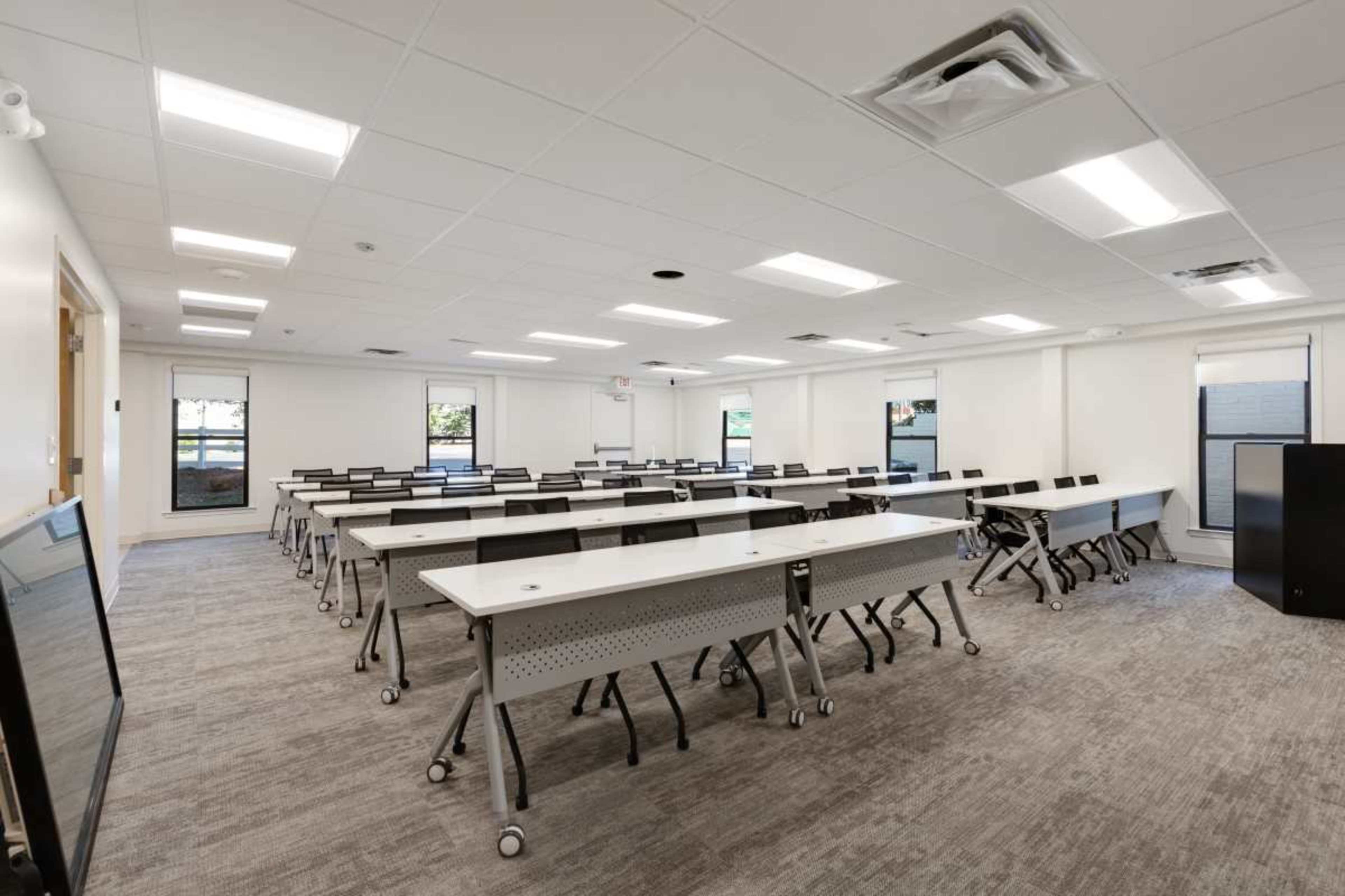The image shows a spacious, brightly lit classroom arranged with rows of movable desks and chairs, featuring large windows and neutral-colored walls.