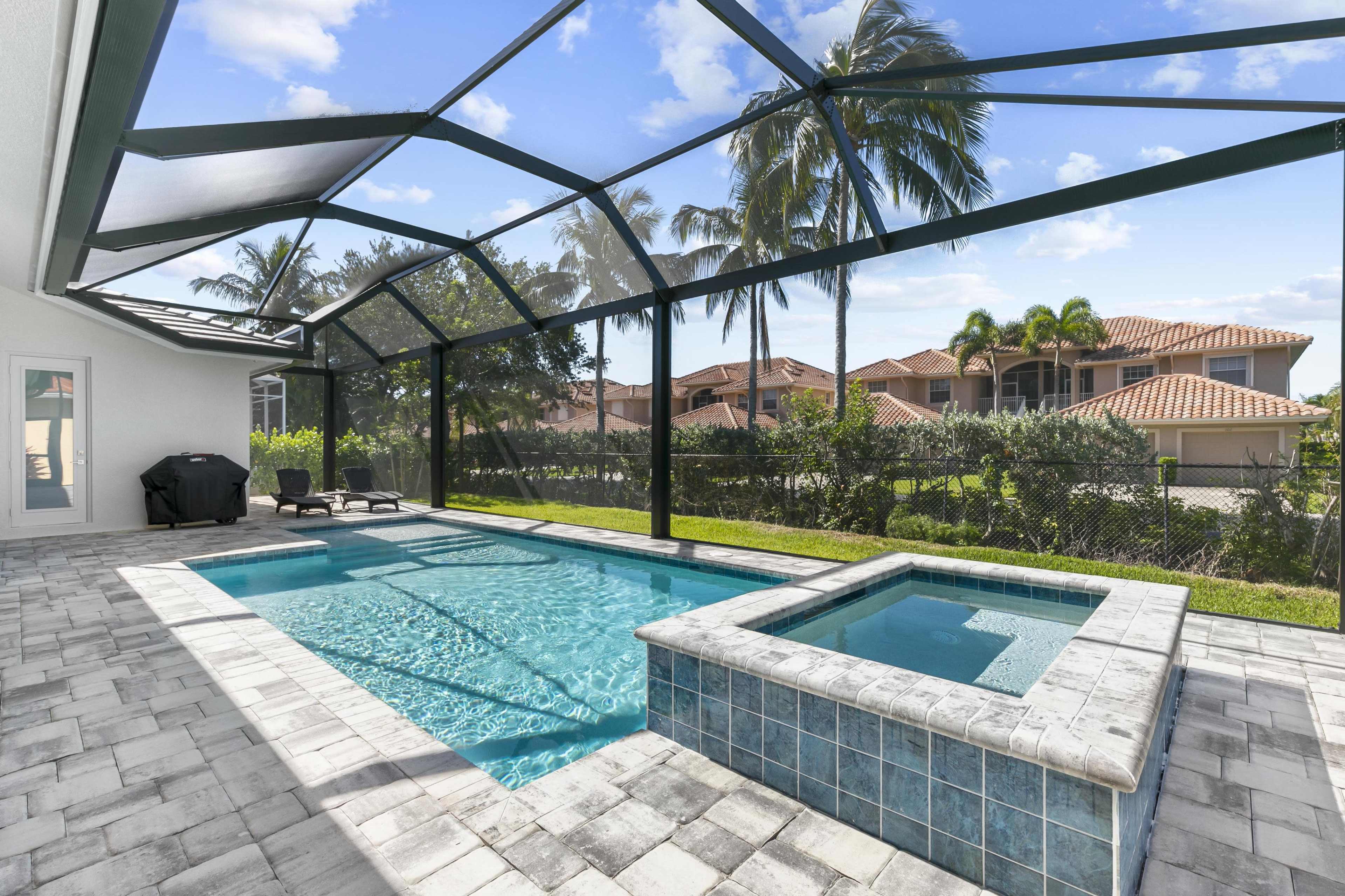 A residential backyard with a swimming pool and a hot tub, enclosed by a glass screen, surrounded by palm trees and houses in the background.