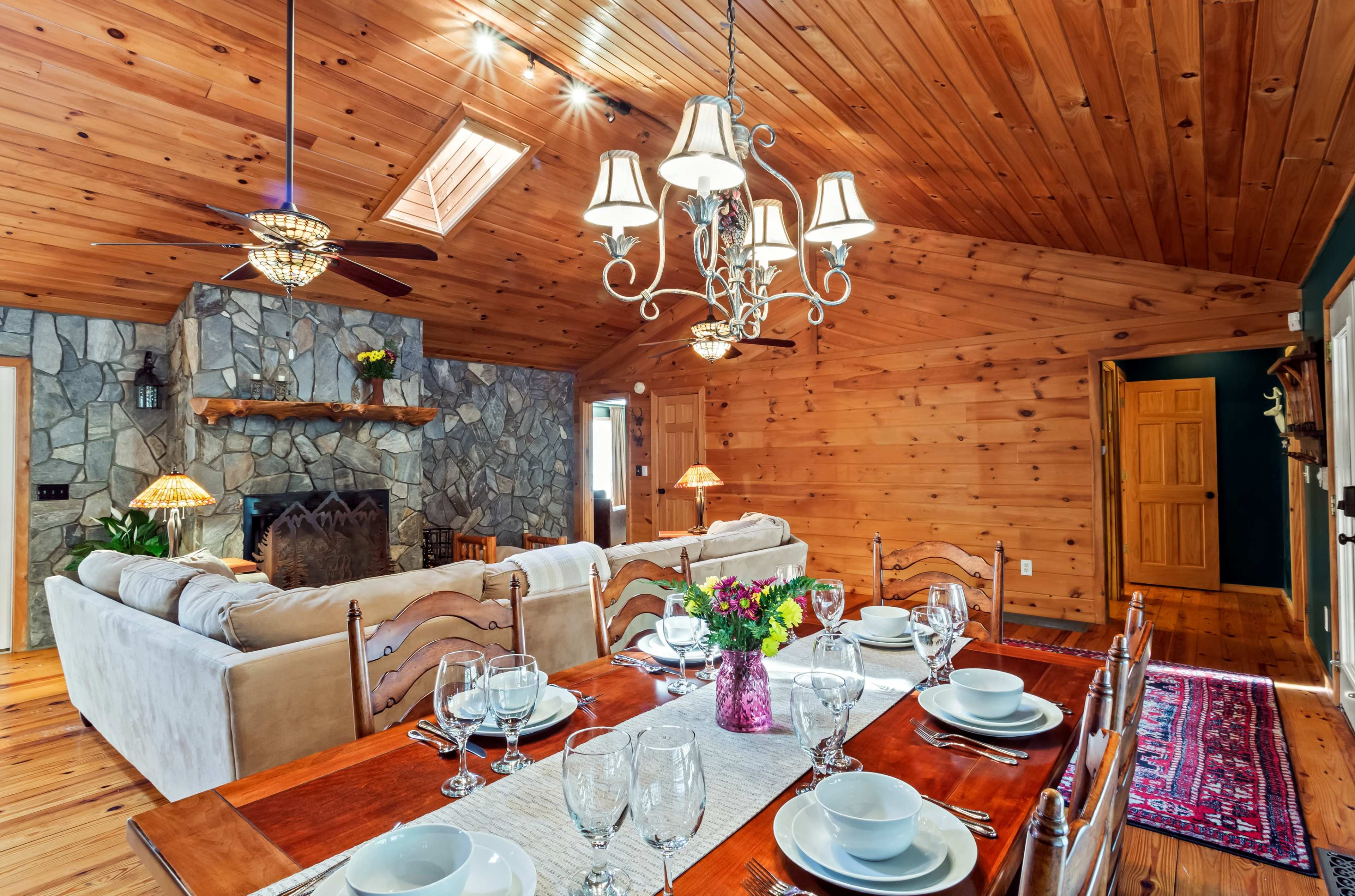 The image showcases a warm, wooden-paneled dining room with a large table set for a meal, a stone fireplace, and a cozy seating area.