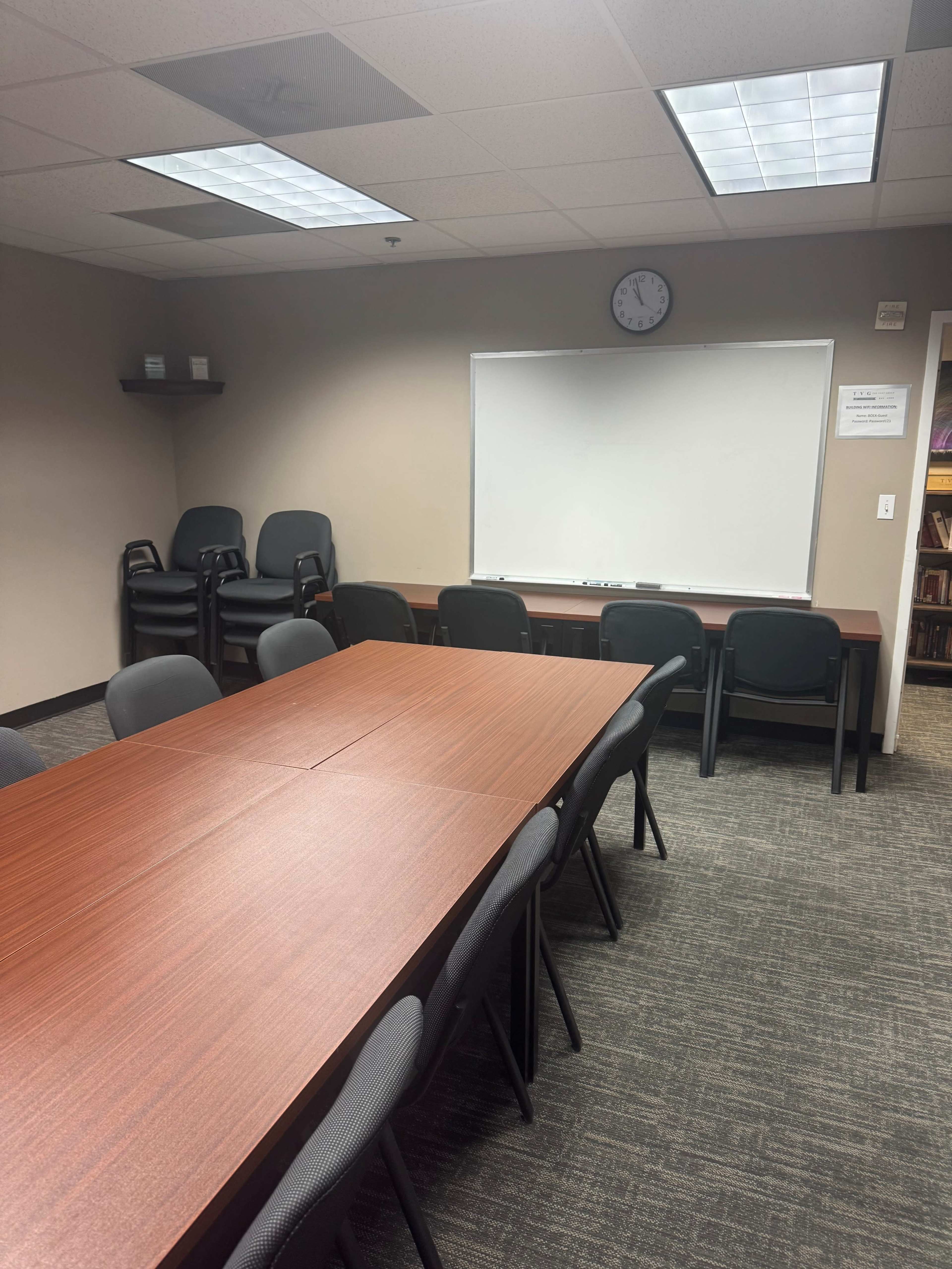The image shows a meeting room with a rectangular table surrounded by black chairs, a whiteboard on the wall, and a clock above it.