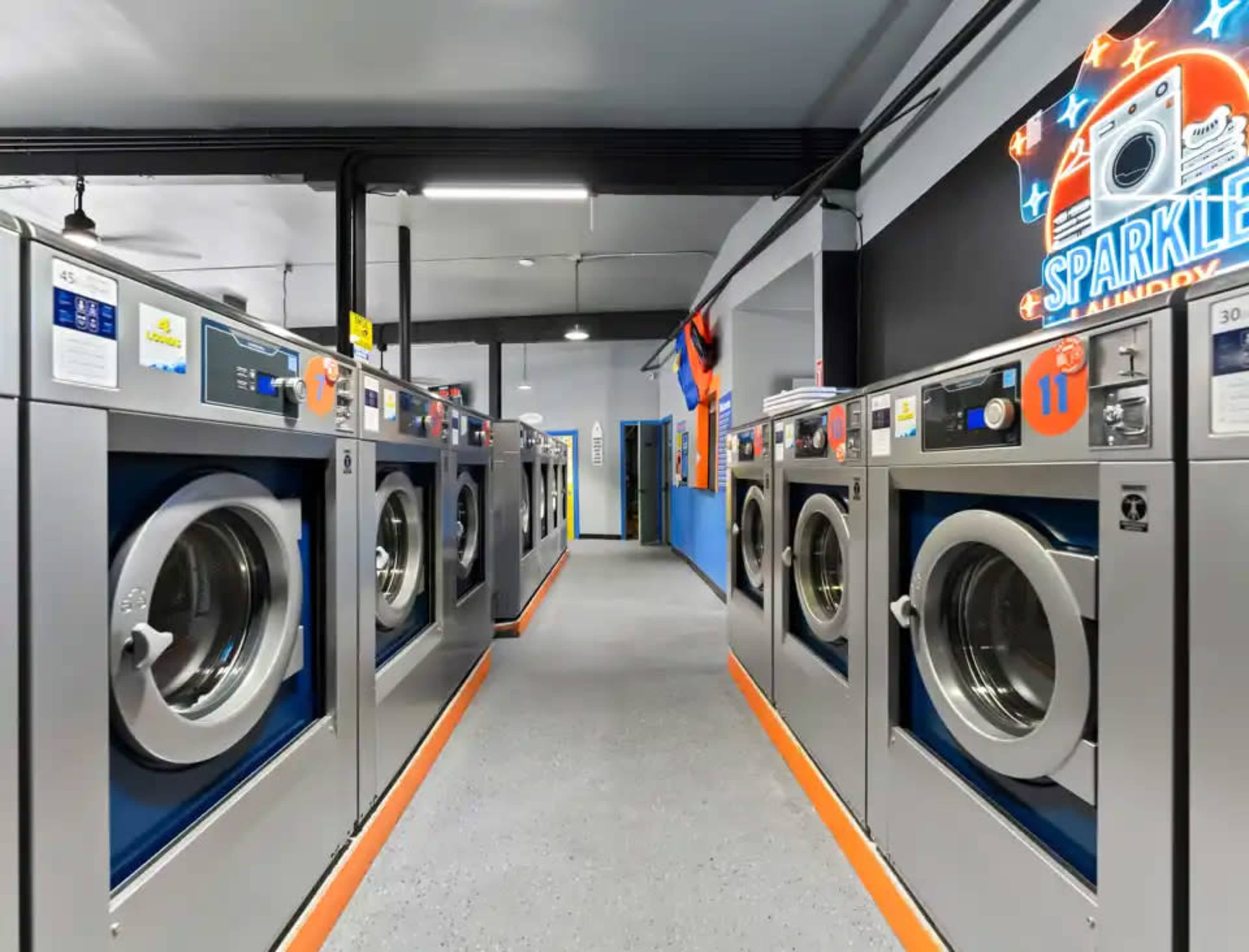 The image shows a modern laundromat with rows of washing machines lined up against a brightly colored wall featuring a logo.