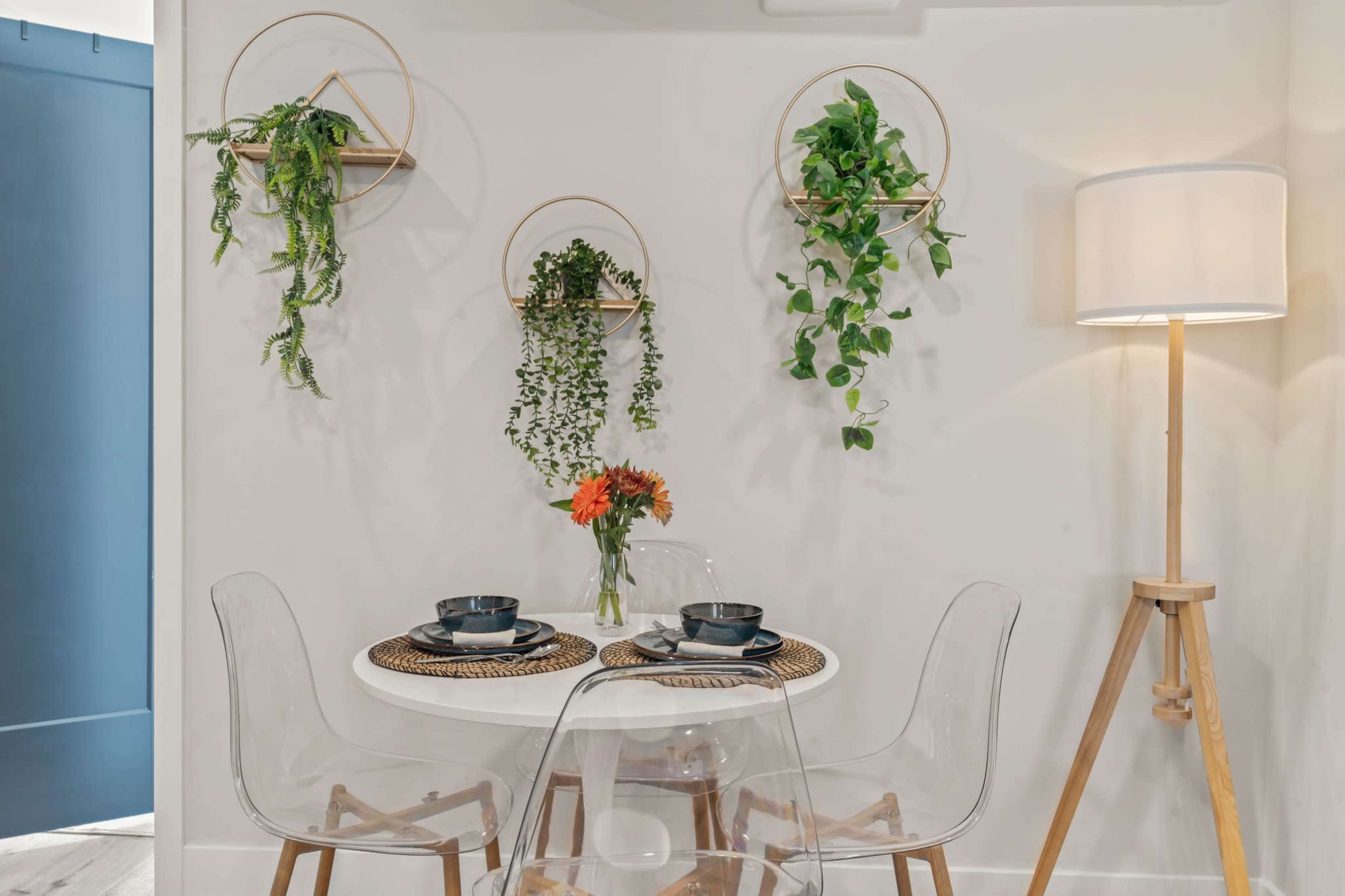 A small dining area features a round table set for two with clear plastic chairs, surrounded by wall-mounted planters holding various green plants and a standing lamp.