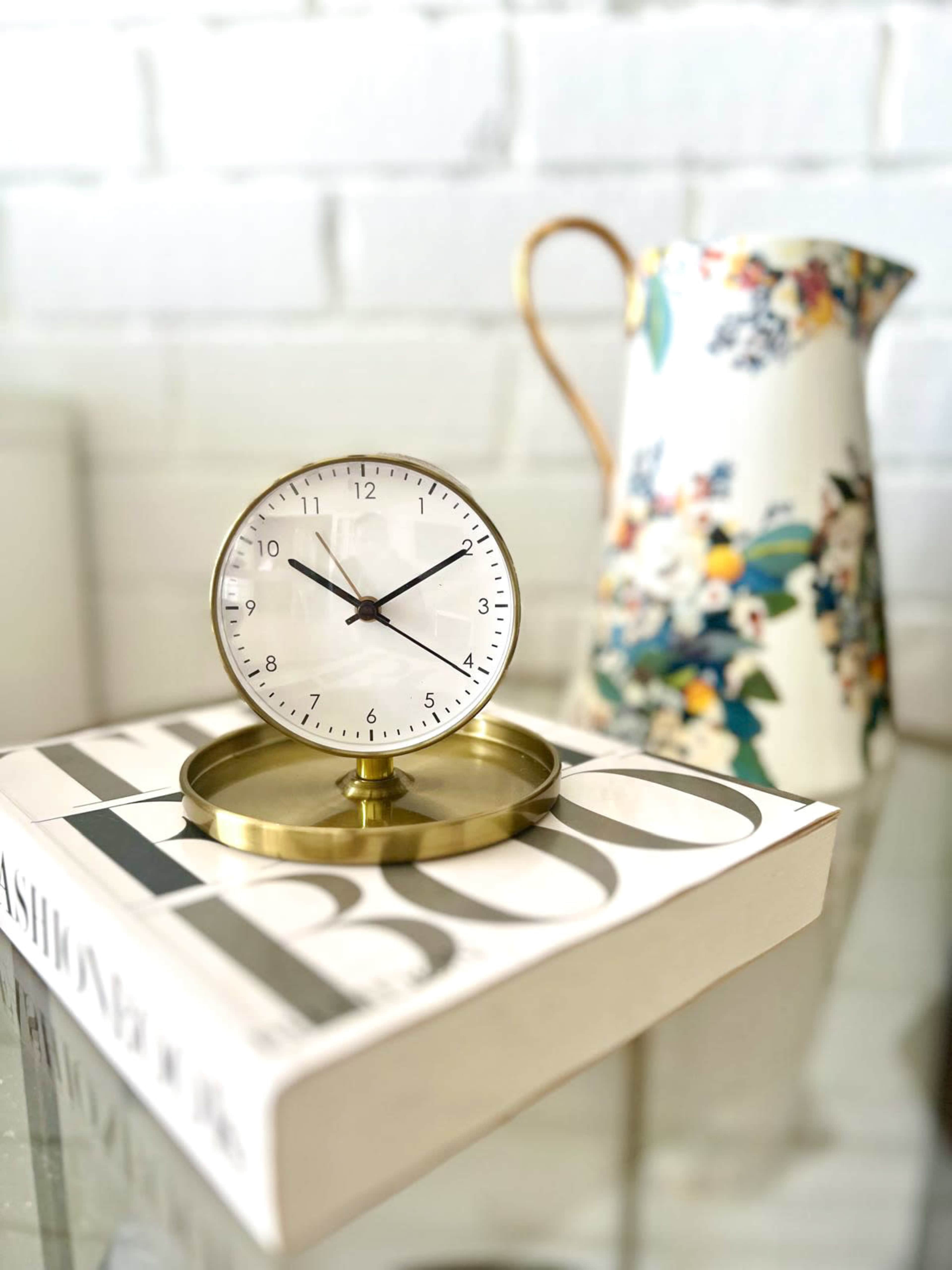 A brass clock sits atop a white book with "Fashion" printed on the cover, next to a floral-patterned pitcher against a brick wall.
