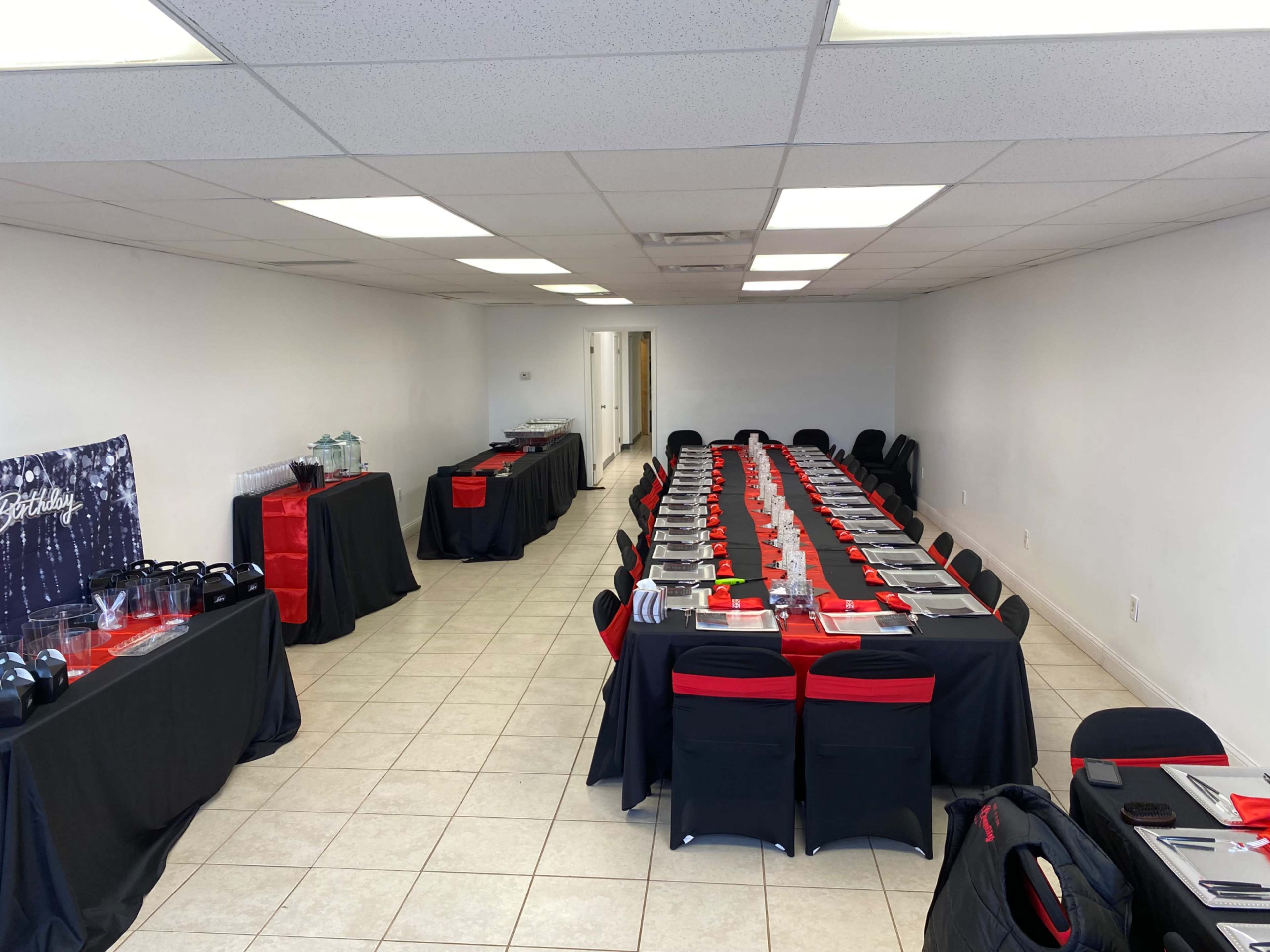 The image shows a neatly arranged banquet hall with a long table set for an event, featuring black tablecloths and red accents, along with a drink station along one wall.