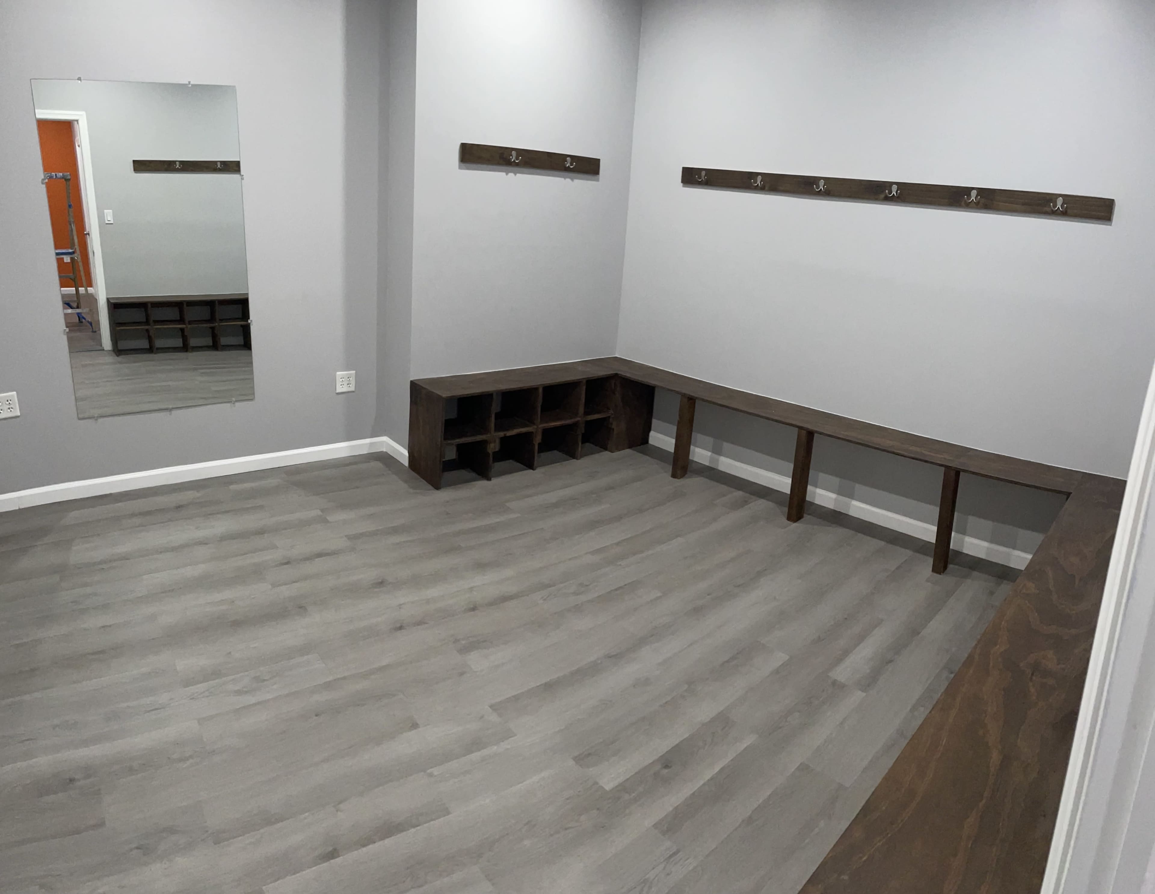 The image shows a minimalist changing room with a mirror, wooden bench, hooks on the wall, and a gray floor.