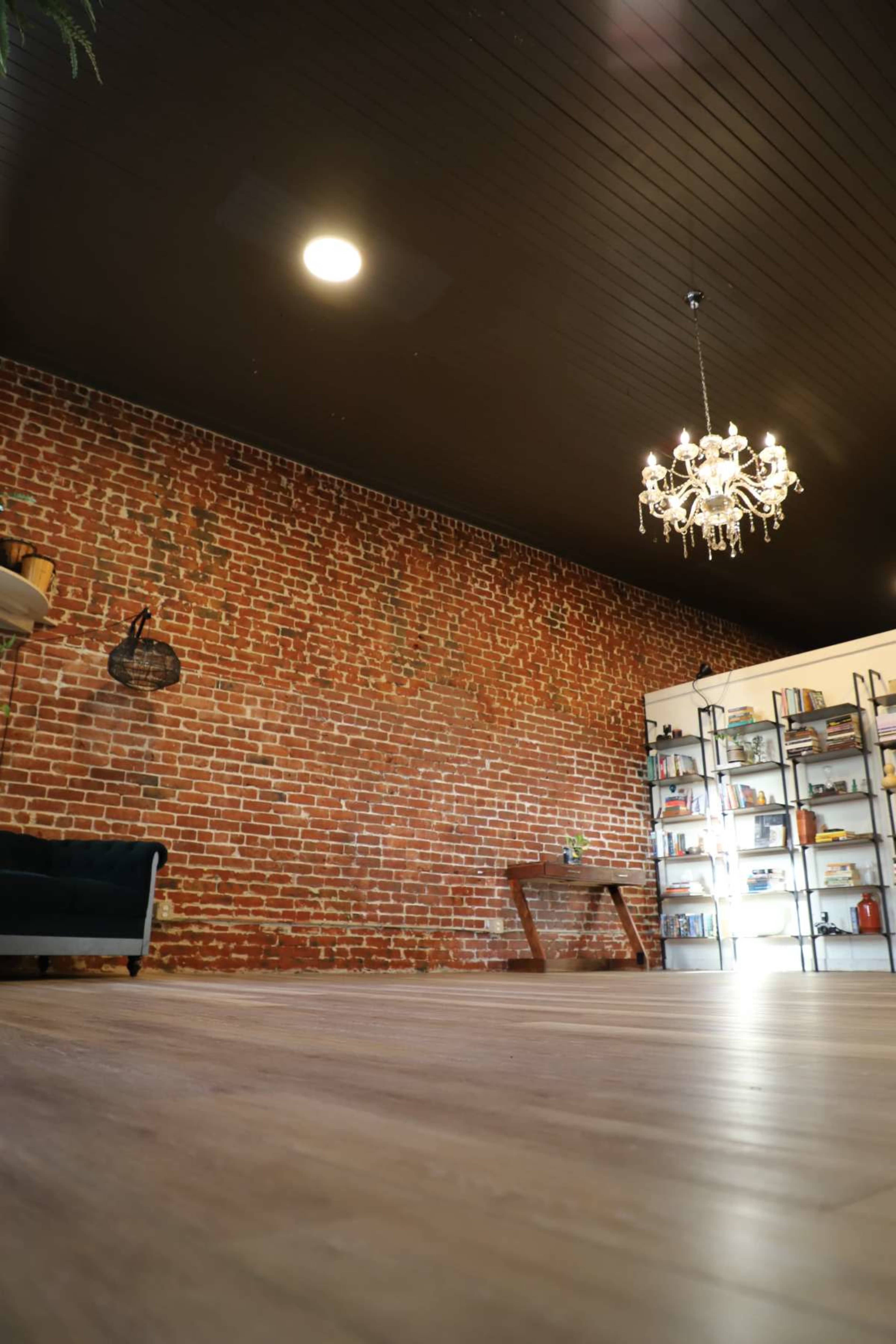 The image shows an interior space with a brick wall, wooden flooring, a chandelier, and a shelving unit filled with various items.