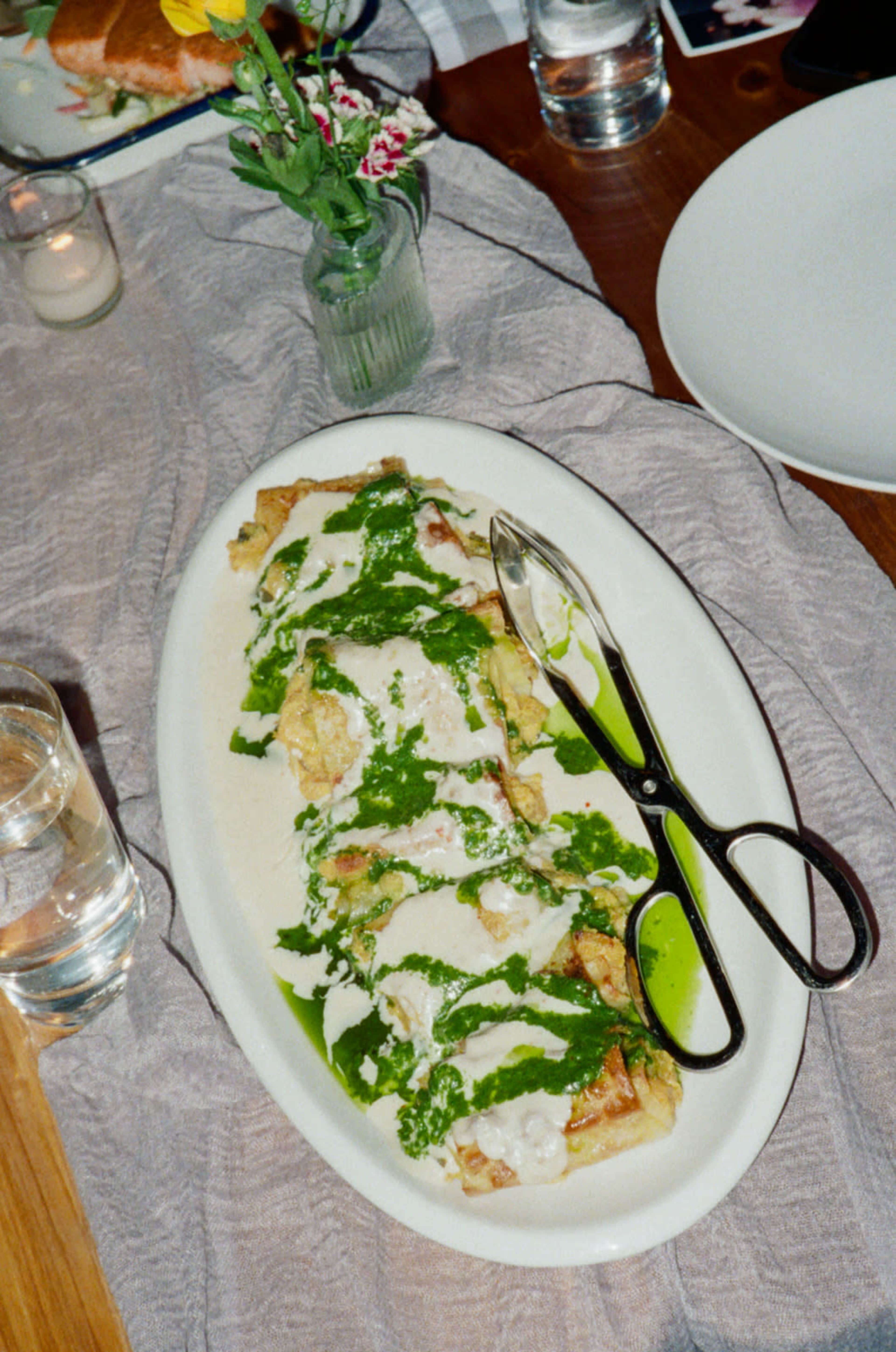 The image features a platter of enchiladas drizzled with green sauce and accompanied by a pair of scissors on a textured tablecloth.