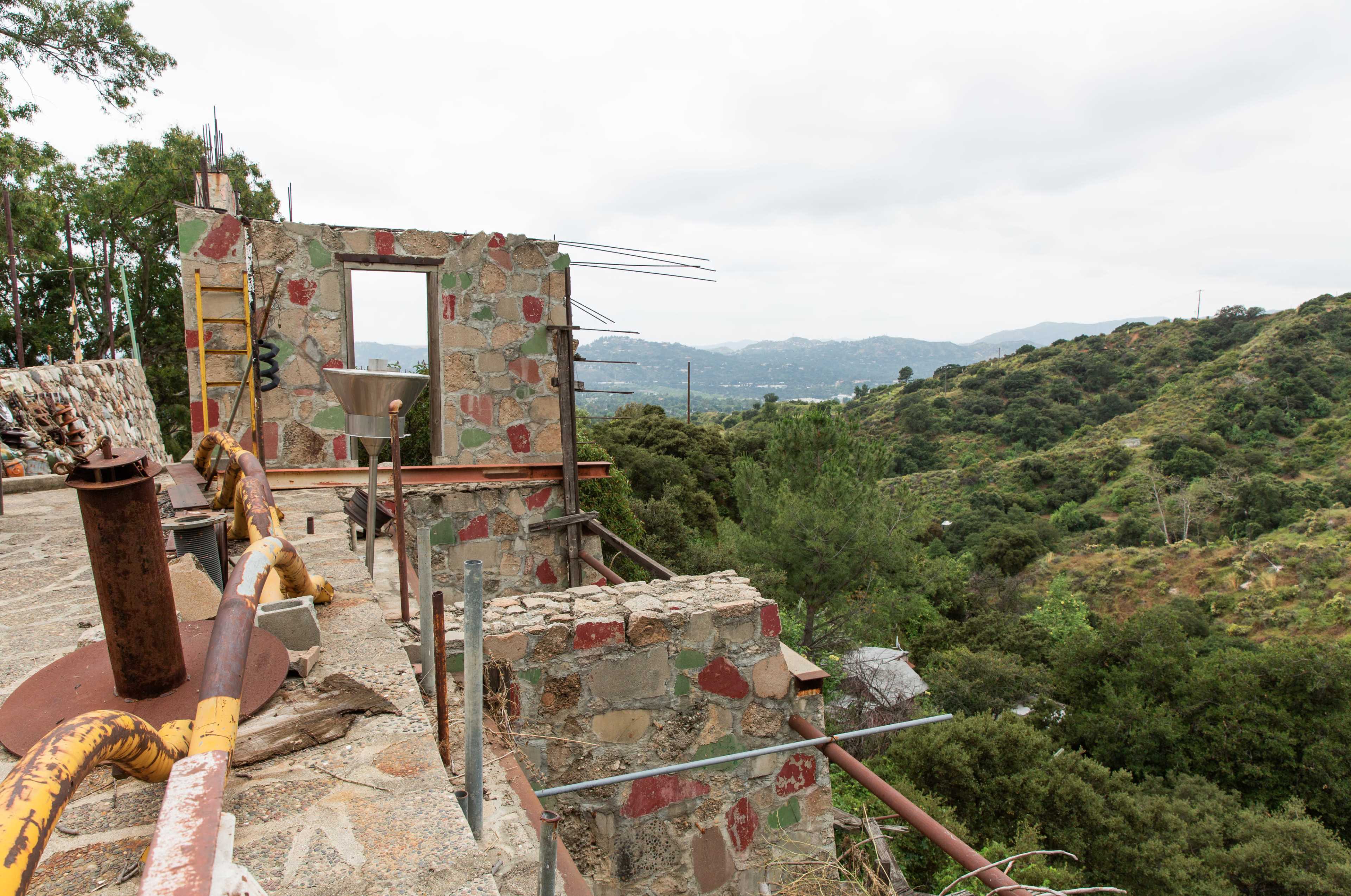 A partially constructed stone building sits on a hillside overlooking a valley and distant mountains under a cloudy sky.
