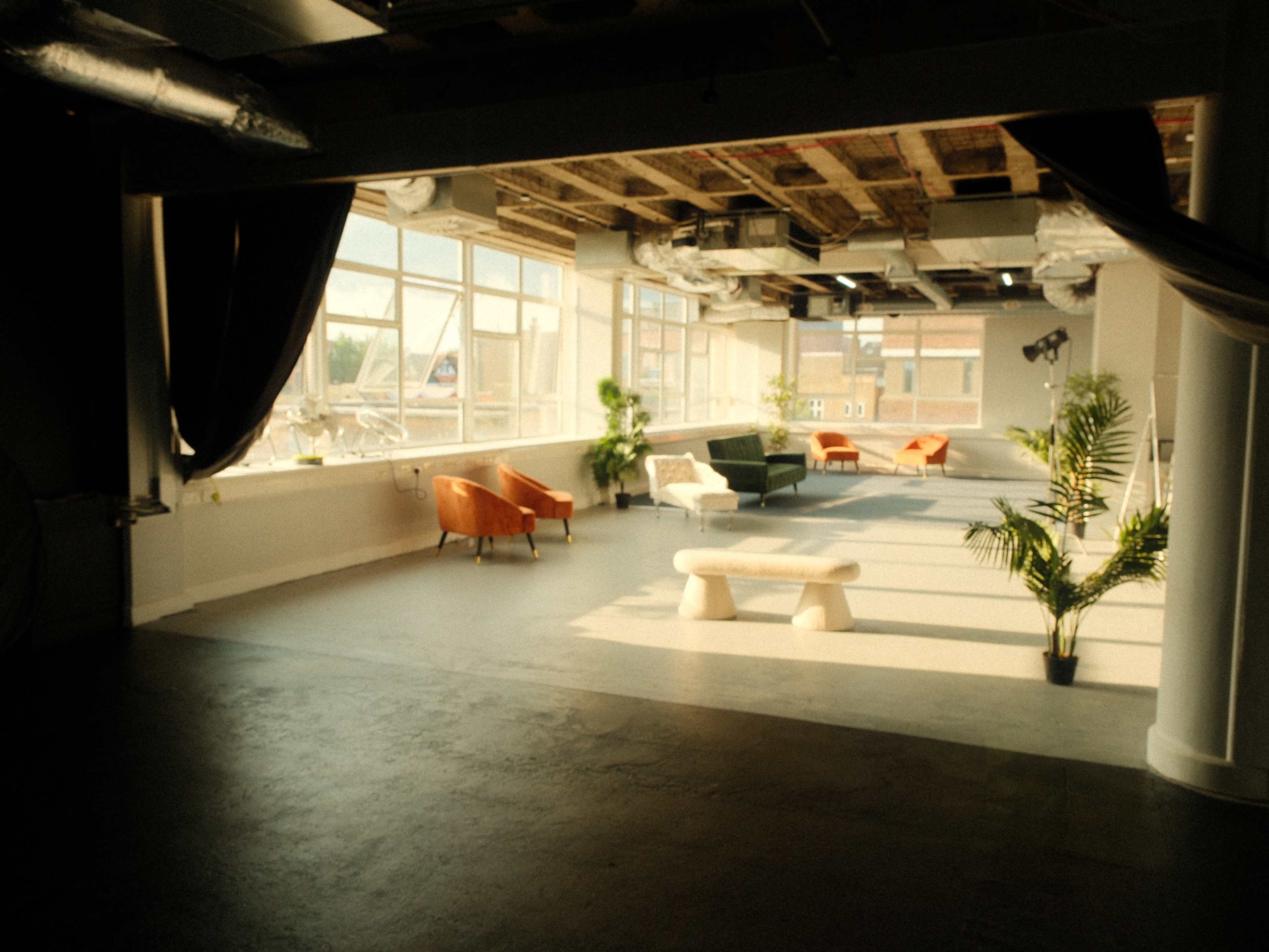 A spacious, modern room features various seating options, including orange and green chairs, large windows, and plants.