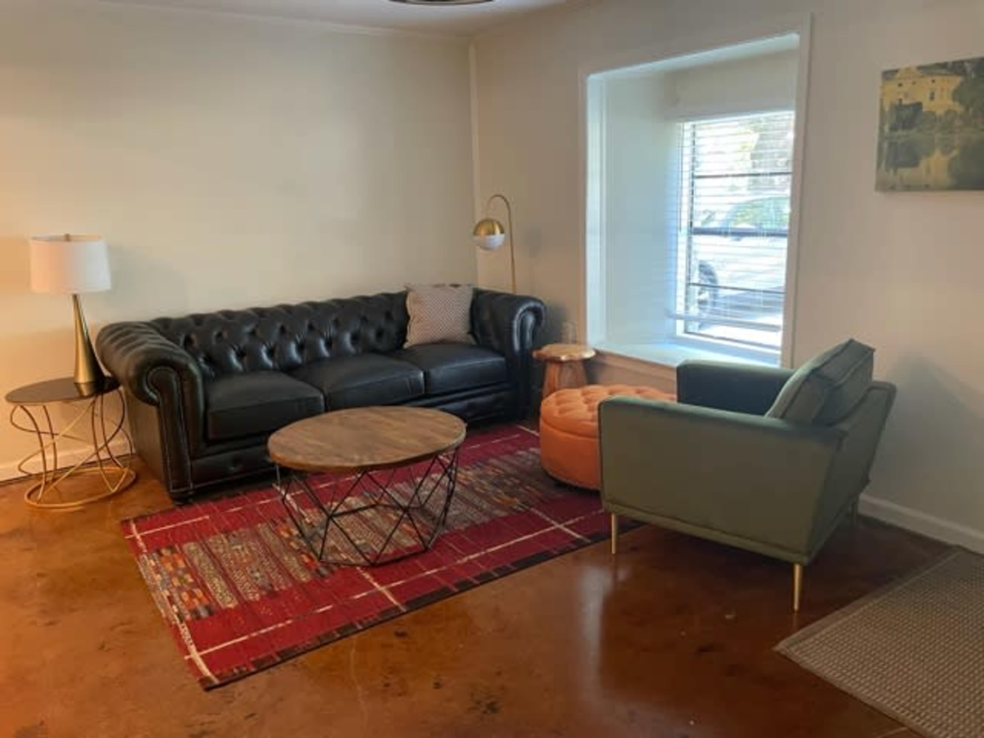 The scene features a stylish living room with a black leather chesterfield sofa, an orange ottoman, a round coffee table, and a green armchair, set on a patterned area rug.
