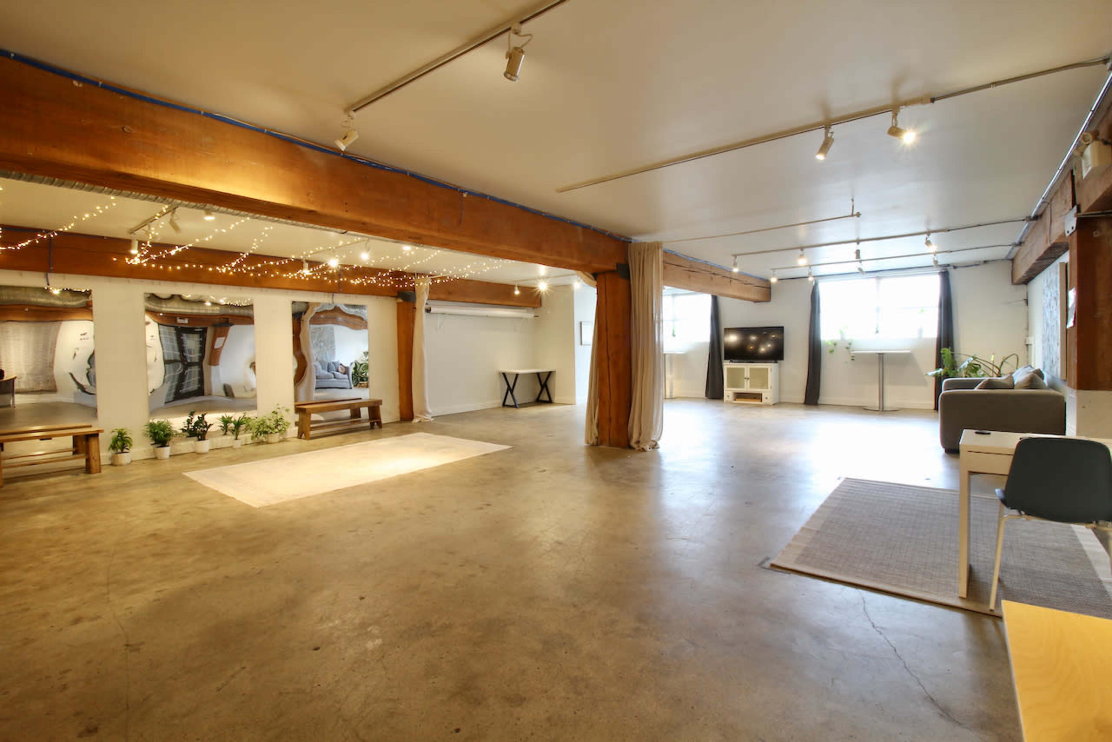 The image shows a spacious, empty indoor venue with wooden beams, concrete flooring, and minimal furniture, including benches and a TV.
