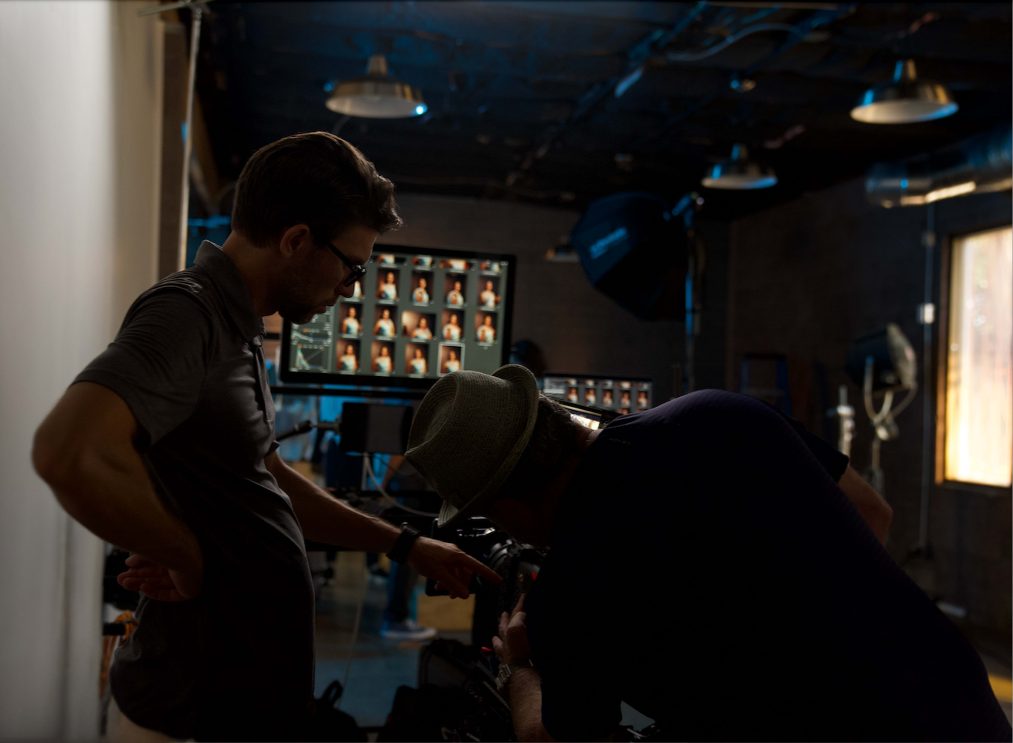 Two individuals are working together in a dimly lit studio, focusing on a camera setup while a monitor displays multiple images.