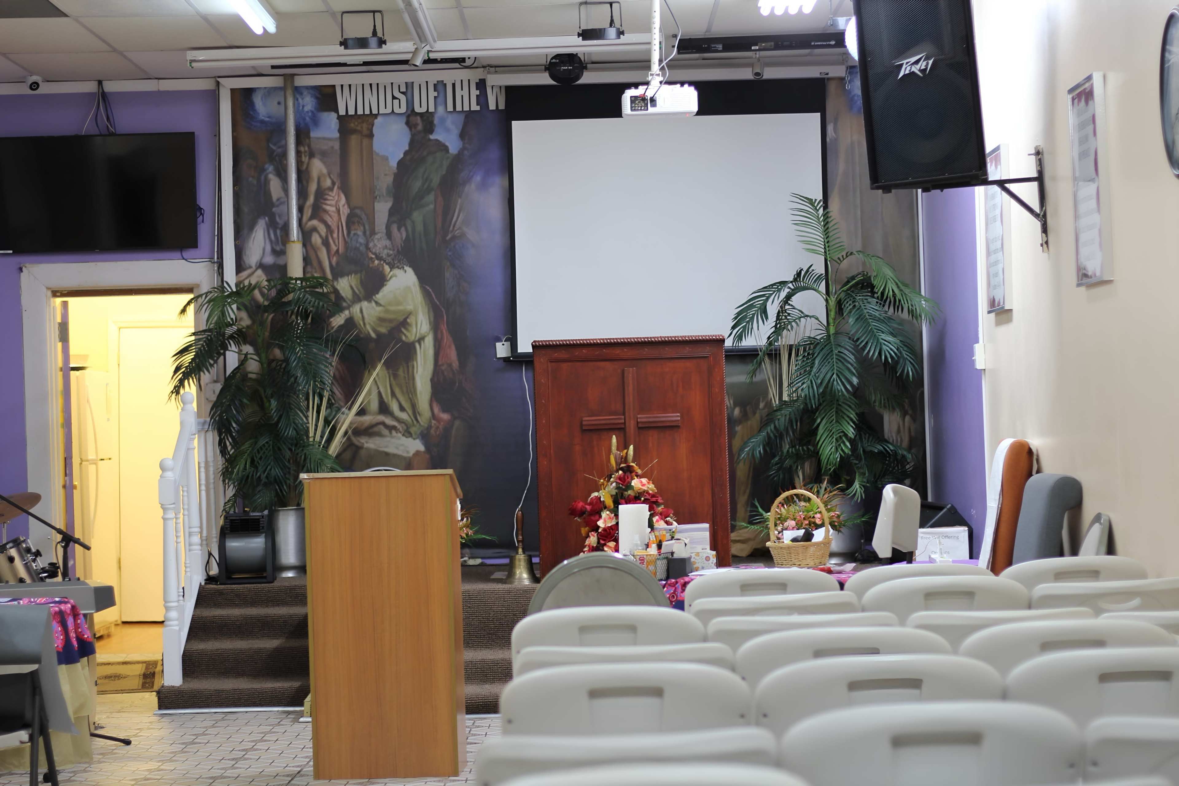 The image shows the interior of a small church or meeting room featuring rows of white chairs, a wooden podium, and a wooden pulpit adorned with plants and decorative items, with a large screen in the background.