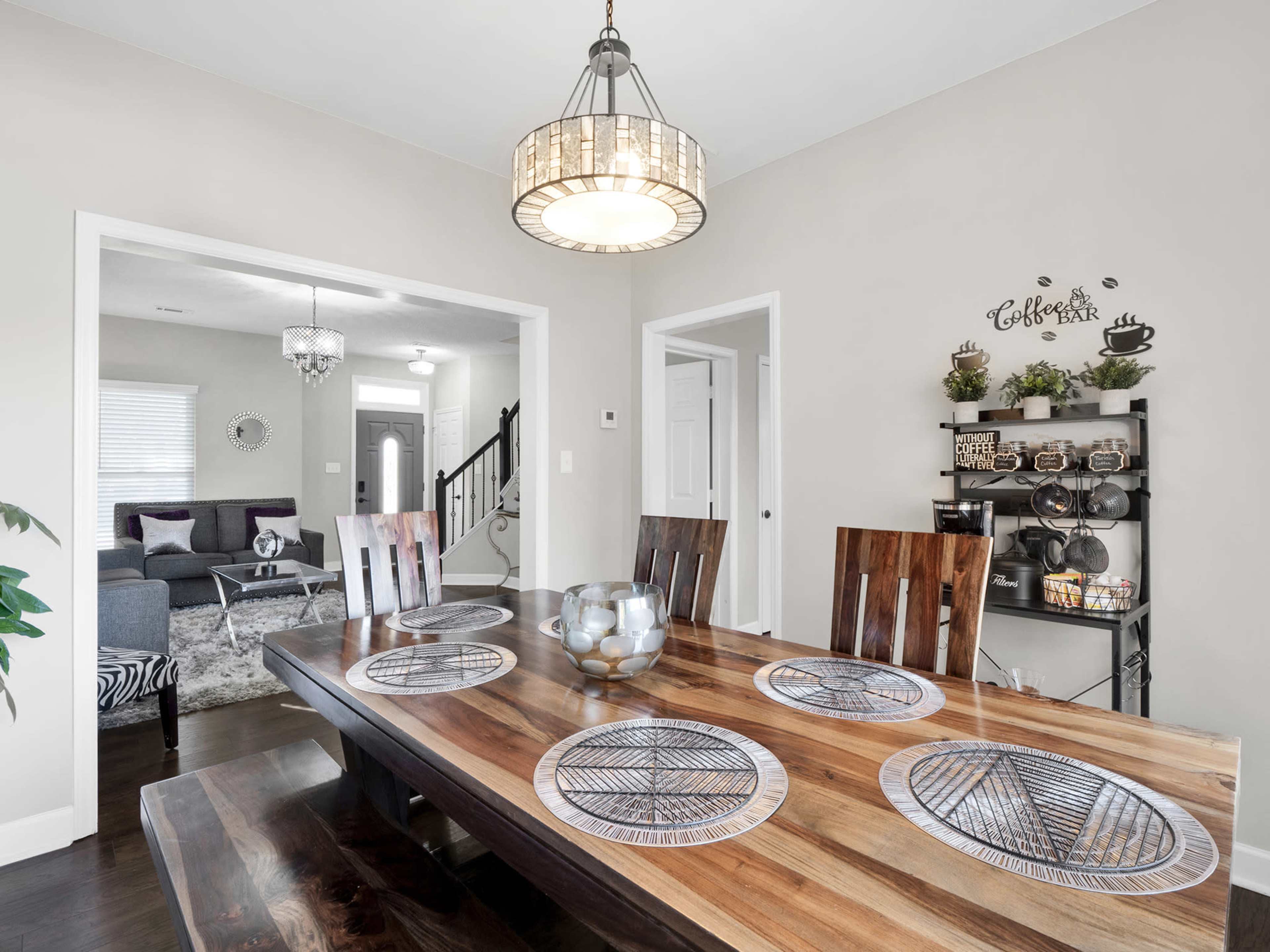 A dining room with a wooden table set for dinner, featuring a decorative centerpiece, surrounded by chairs, and an adjoining living space visible through an open doorway.