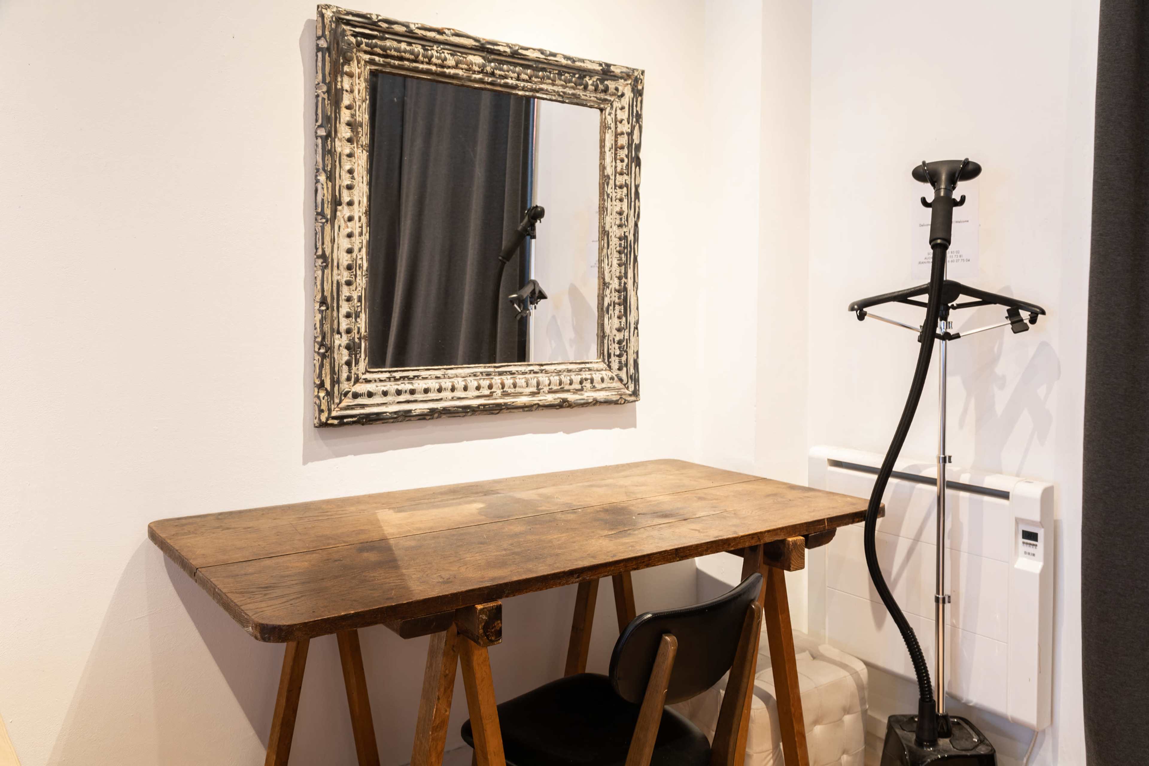 A wooden table with a black chair beside a large ornate mirror in a room with white walls and a garment steamer in the corner.