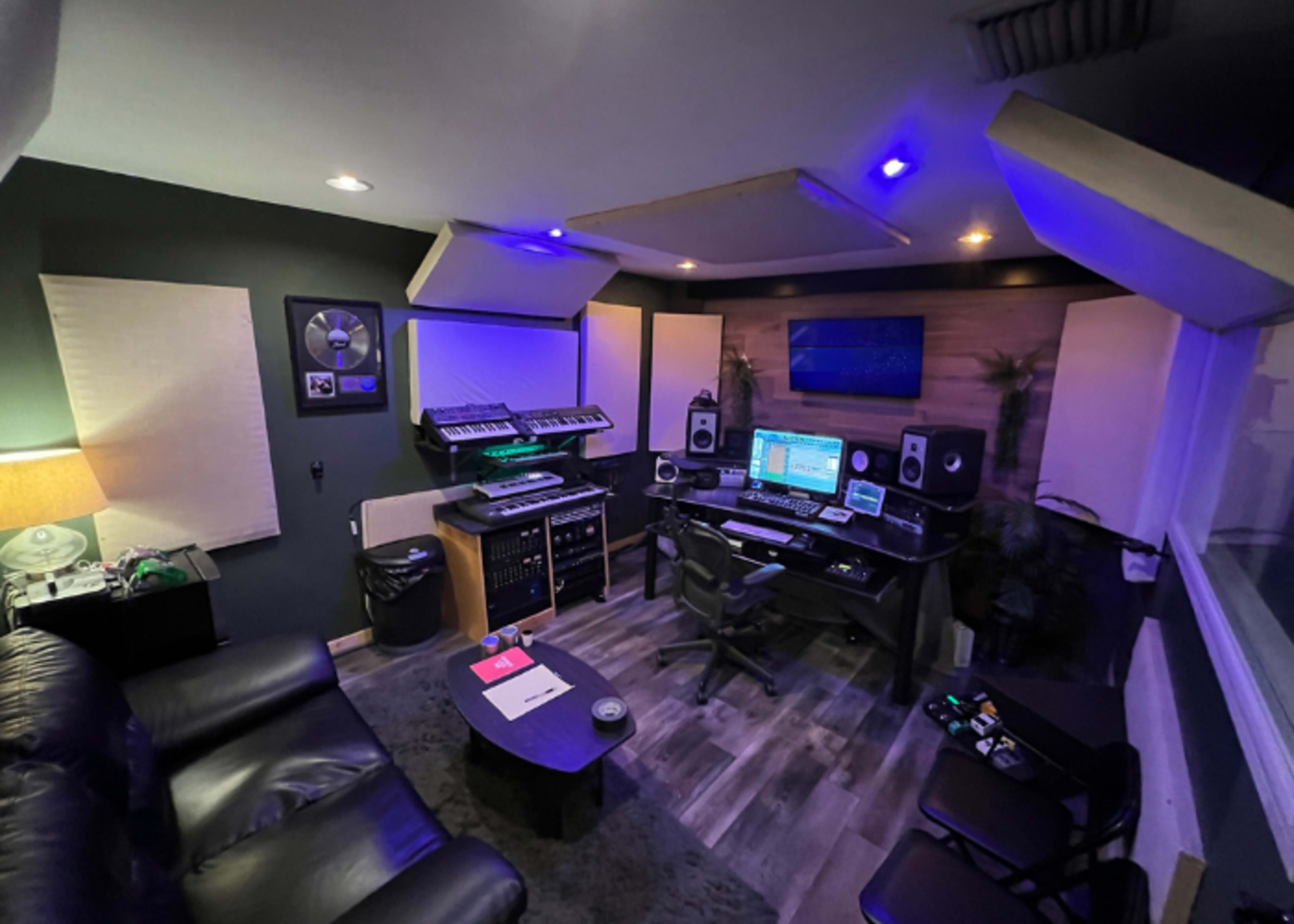 The image shows a small music production studio featuring a desk with computer equipment, synthesizers, monitors, and acoustic panels on the walls.