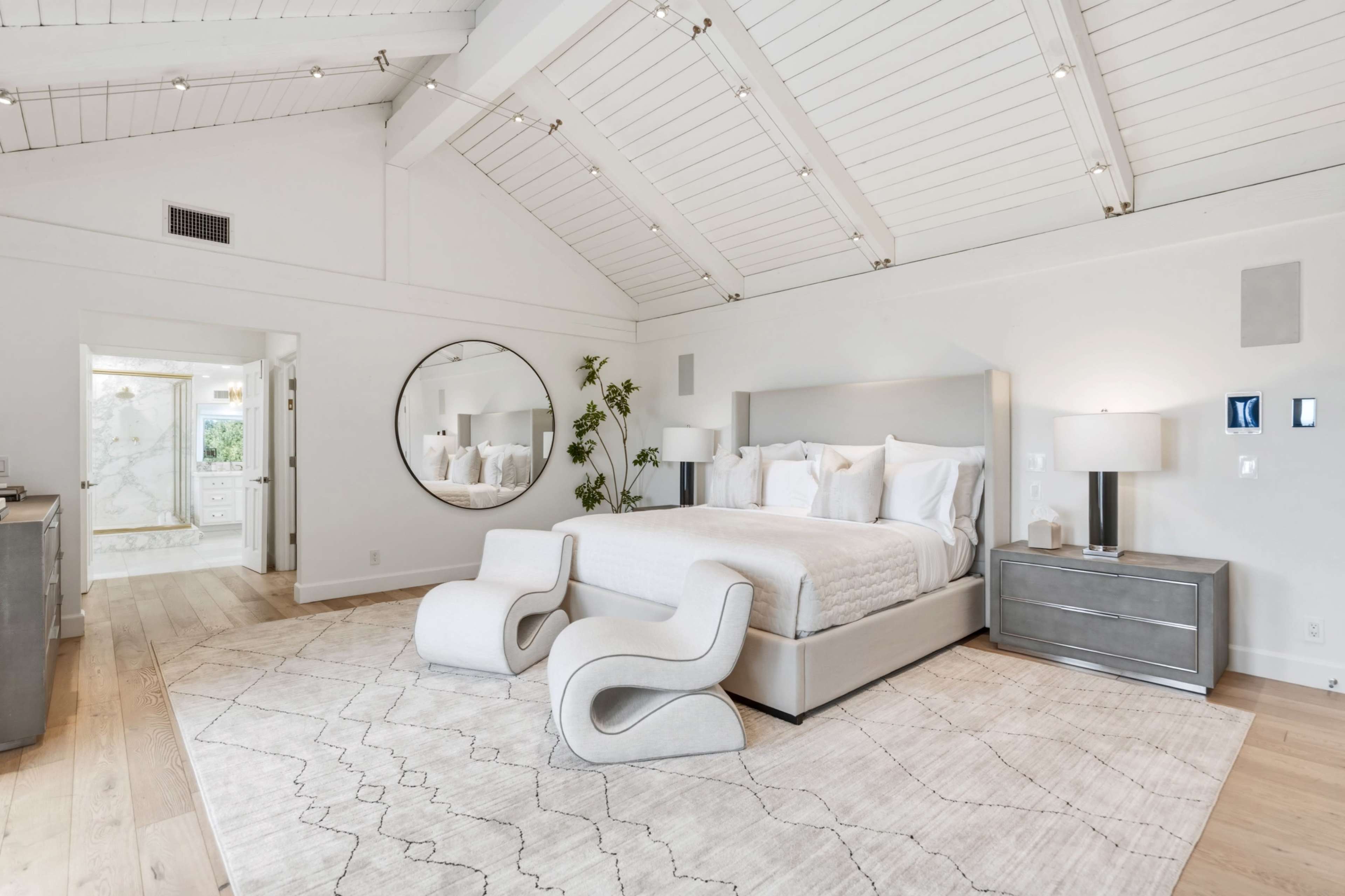 The image shows a modern bedroom with a large bed, circular mirror, two bedside lamps, and a light-colored area rug on a wooden floor.