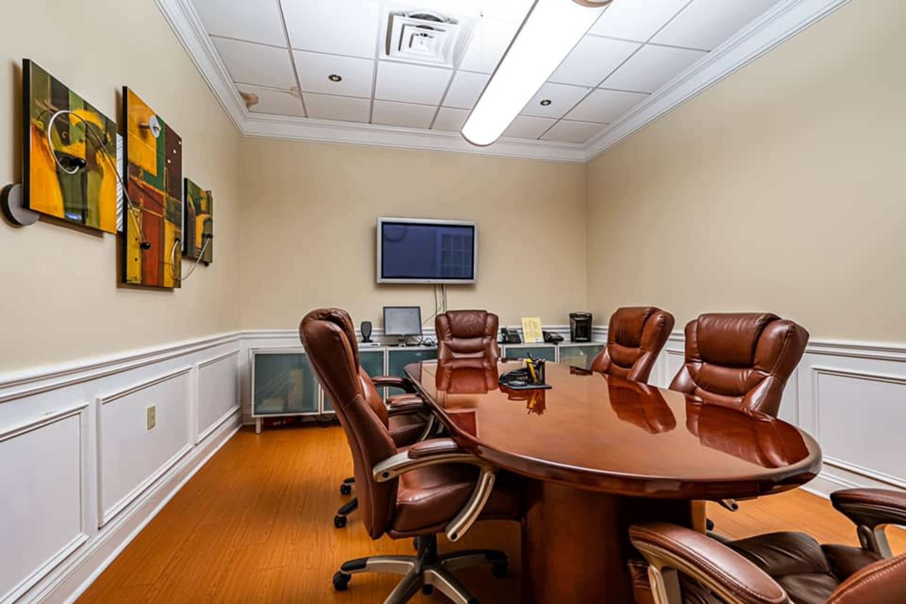 The image shows a professionally arranged conference room featuring a large oval wooden table surrounded by several leather chairs, with a wall-mounted TV and artwork on the walls.