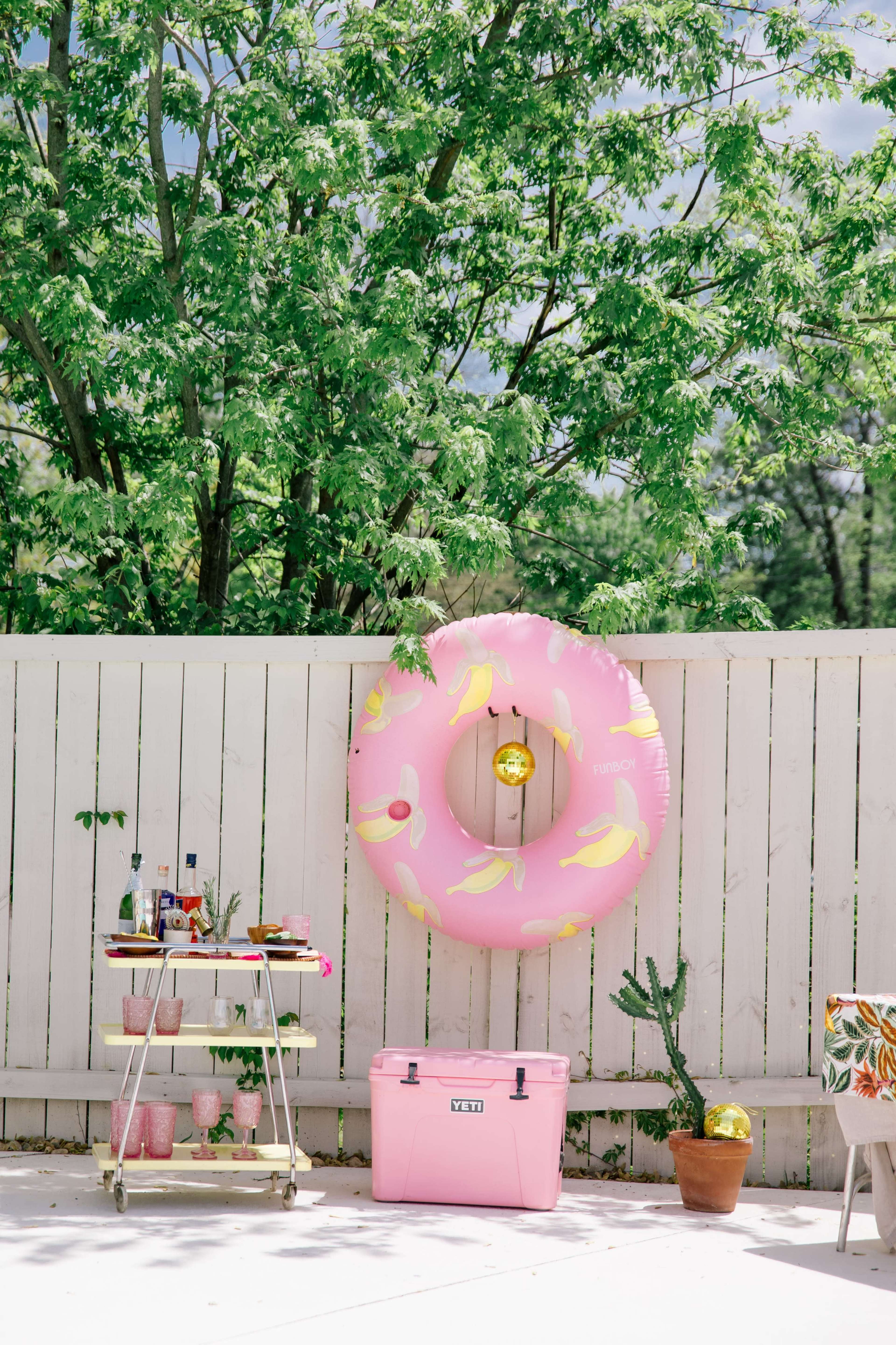 The scene features a pink inflatable donut float hanging on a white fence, with a bar cart and a cooler nearby amidst greenery.