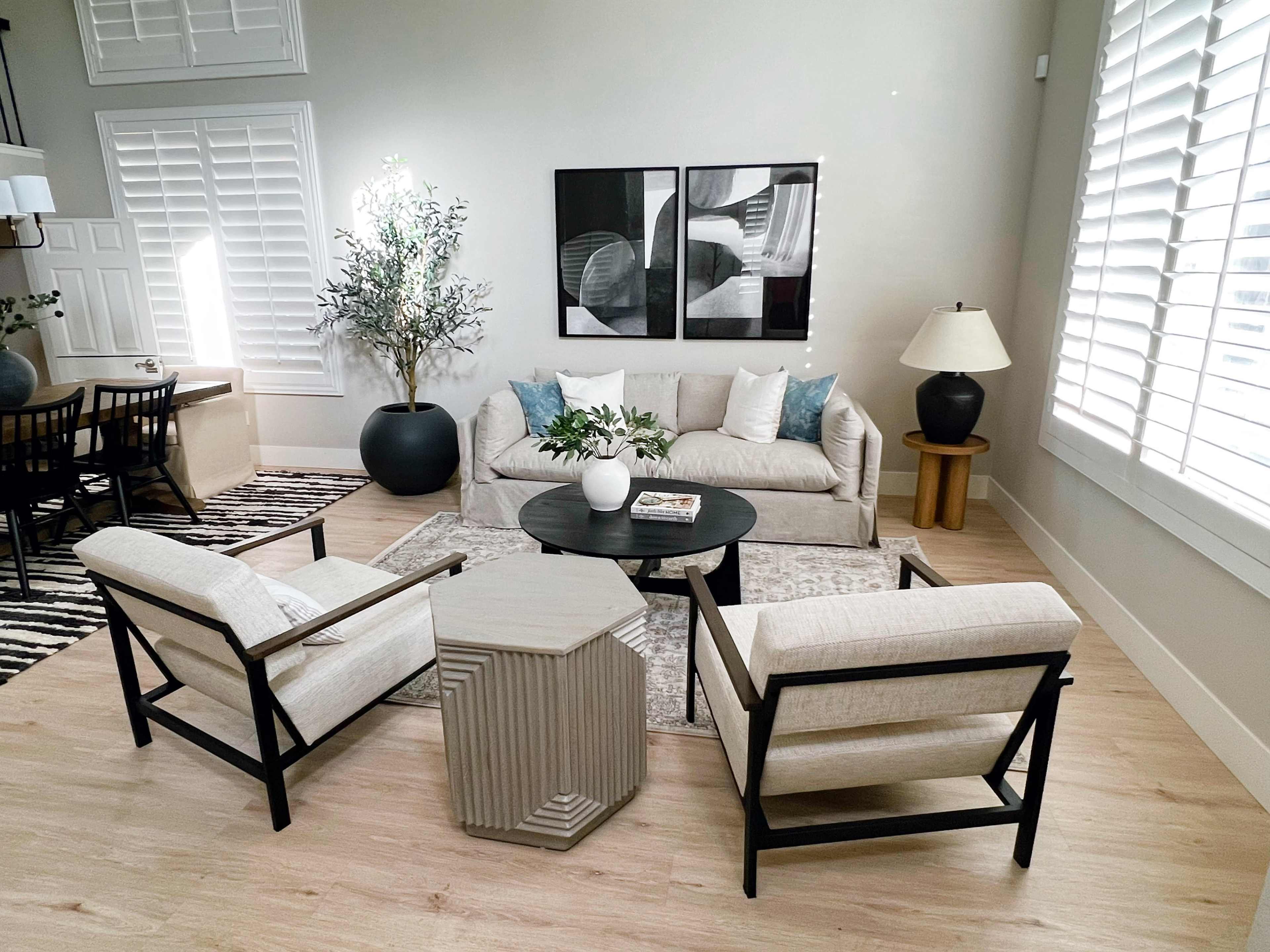 A modern living room with a light-colored sofa, two armchairs, a round coffee table, and a side table, all arranged around a rug and accompanied by decorative plants and artwork on the walls.