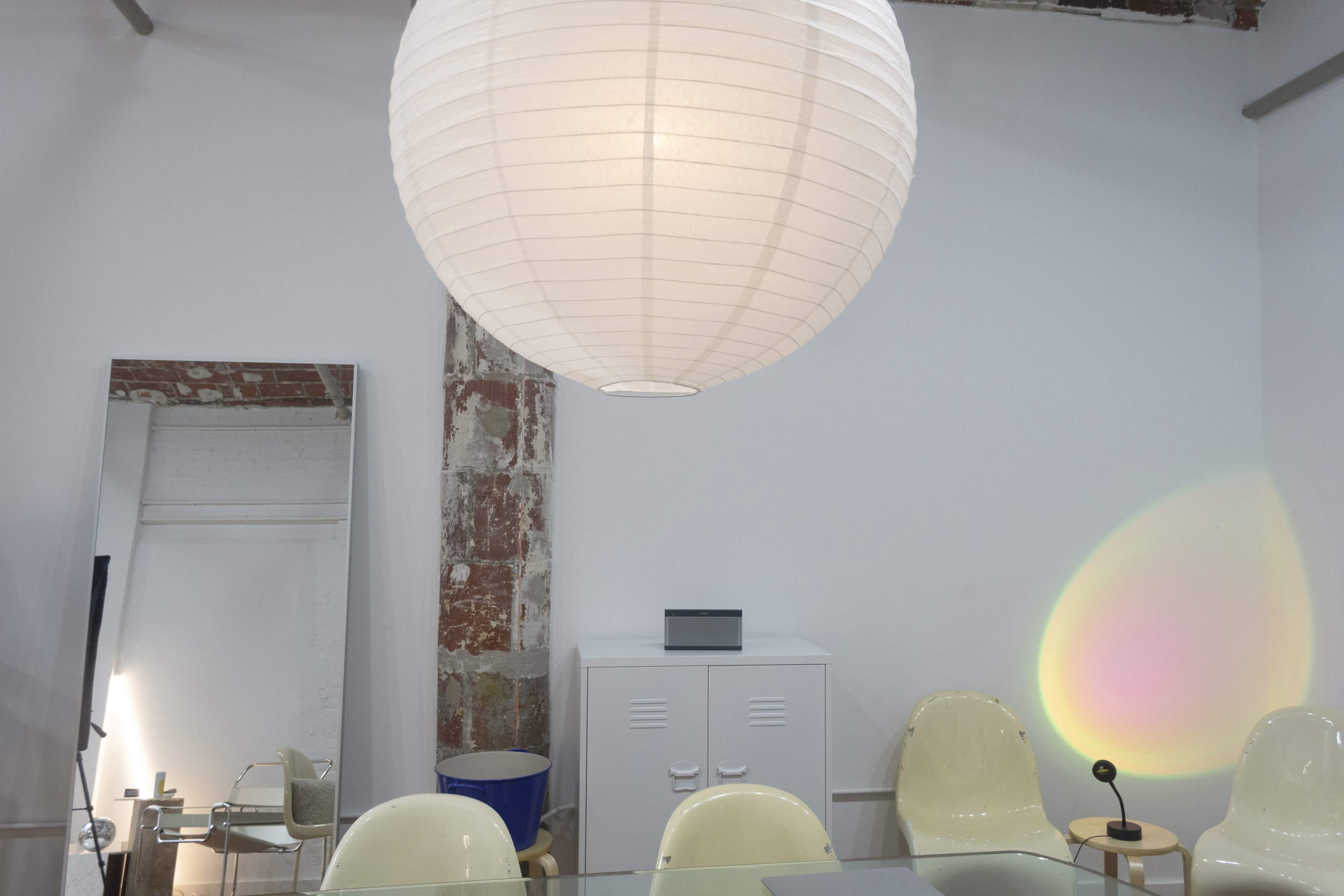 A large paper lantern hangs from the ceiling above a mirrored wall, a white cabinet, and several modern chairs in a minimalist workspace.