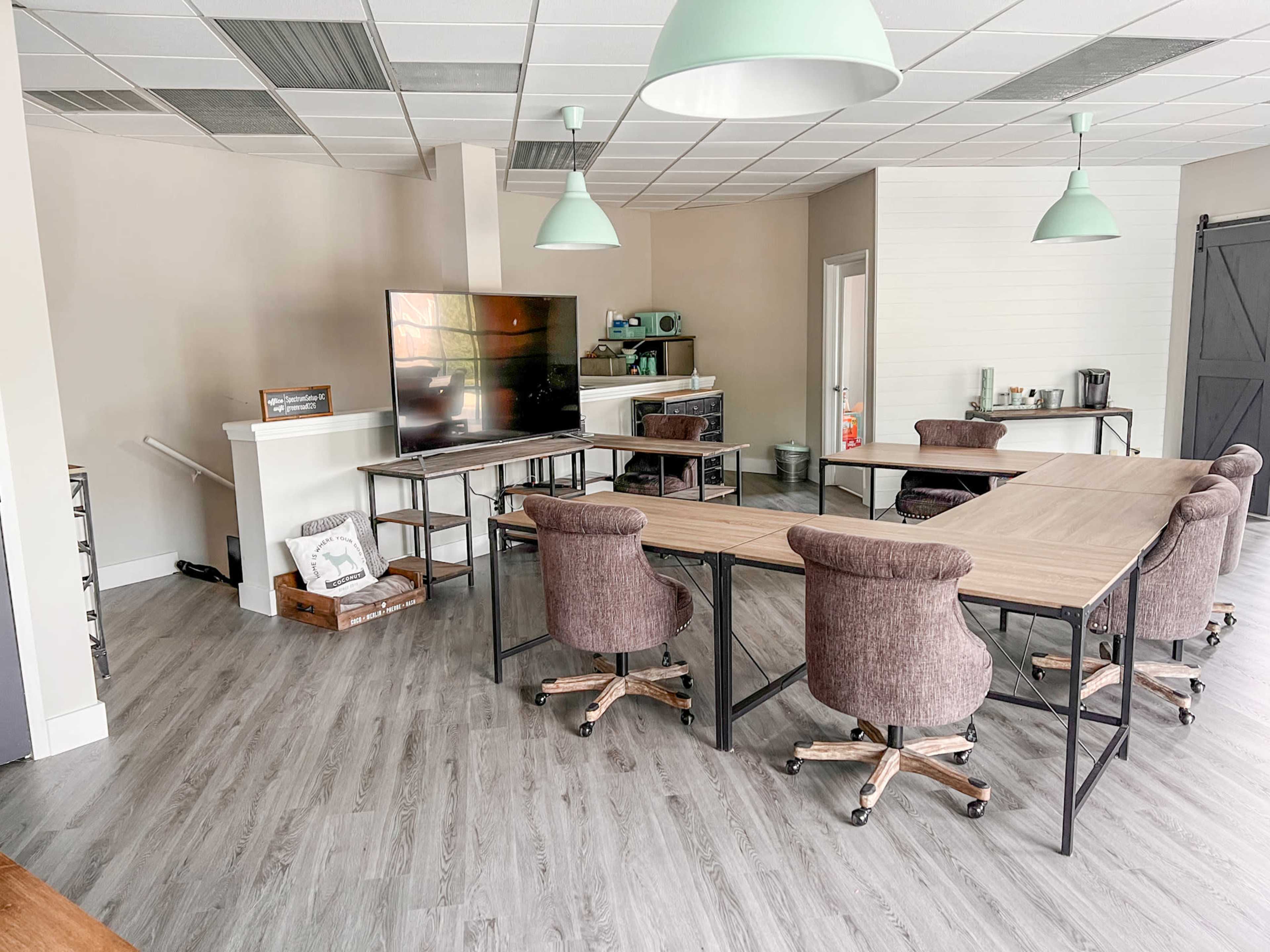 The image shows a modern meeting room with a long rectangular table surrounded by rolling chairs, a television, and a kitchenette in the background.