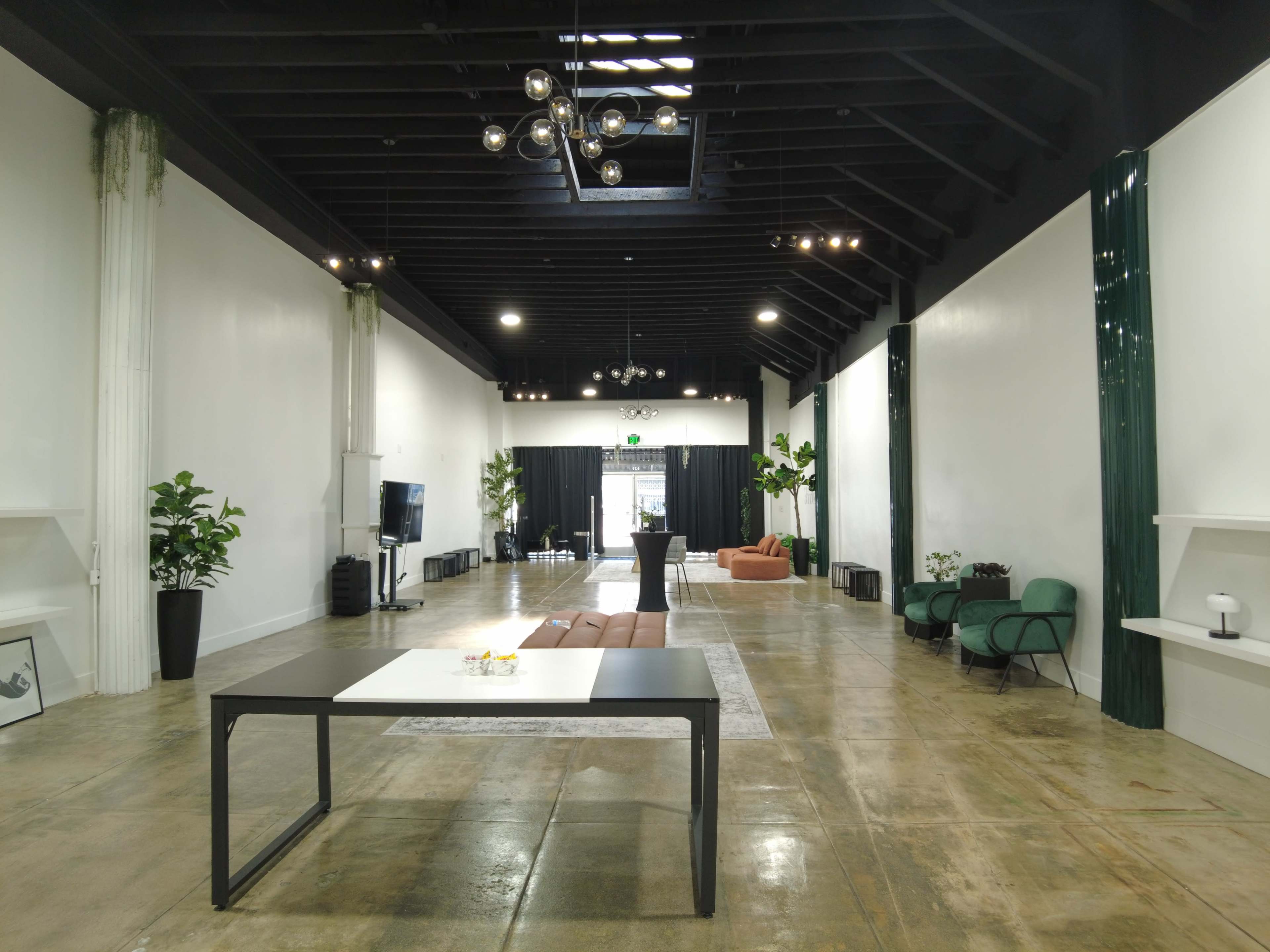 A spacious indoor area features a large table in the center, surrounded by seating areas and plants, with a black curtain at the far end.