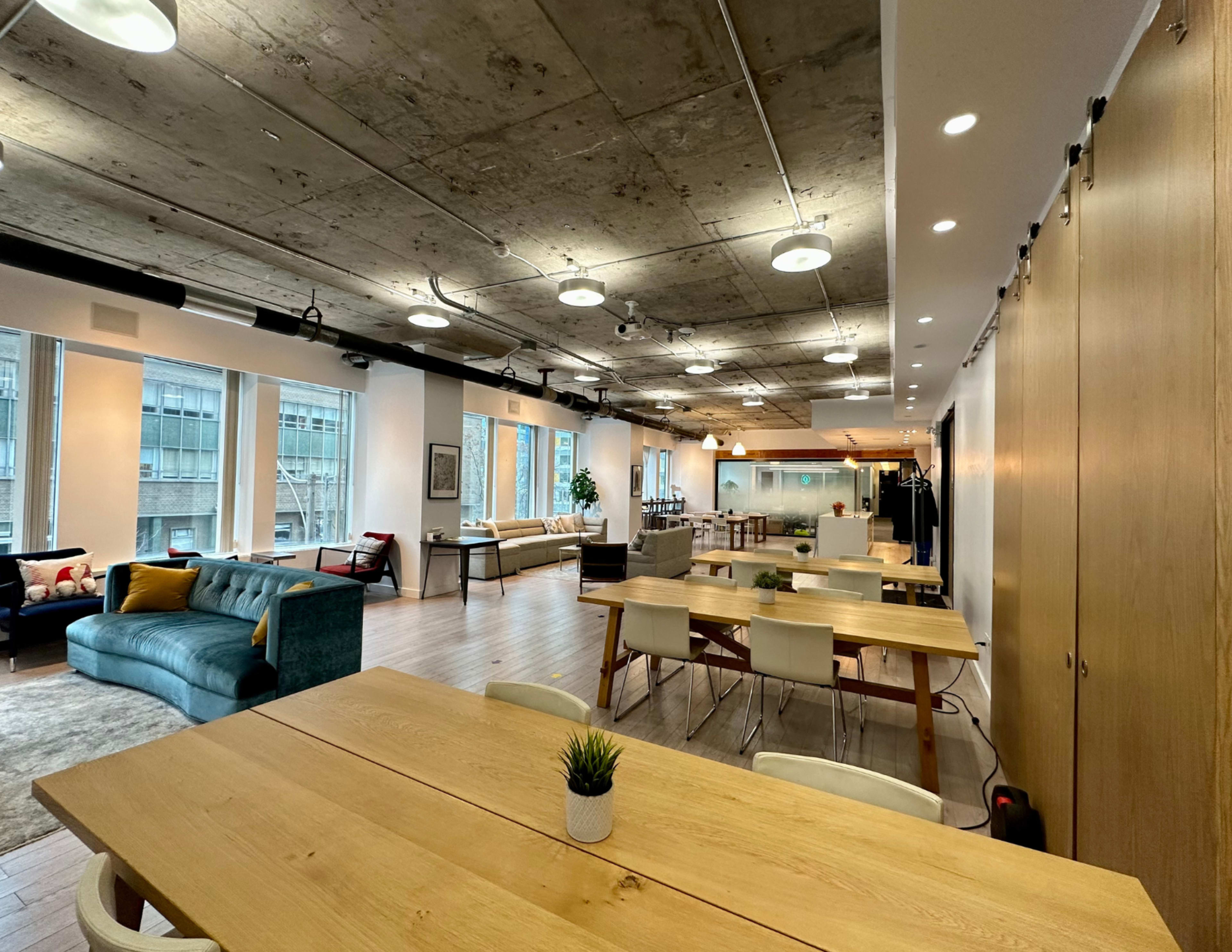 An open office space featuring wooden tables, a couch, and large windows that let in natural light.