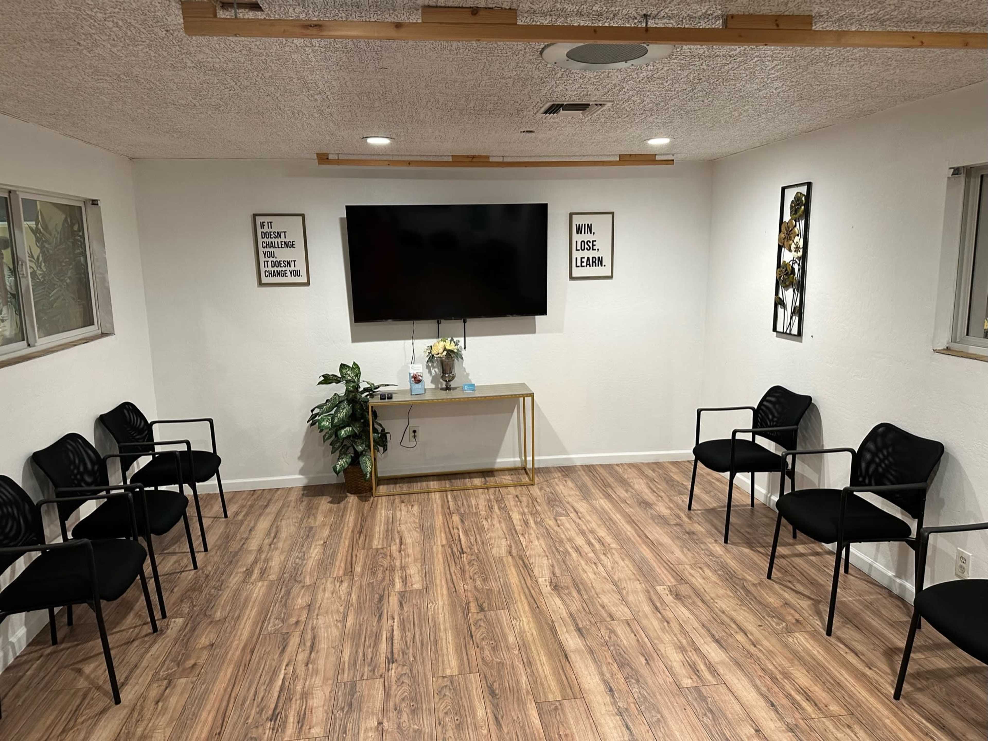 A minimalistic room with a large wall-mounted TV, a table, several black chairs arranged in a circle, and two motivational posters on the walls.