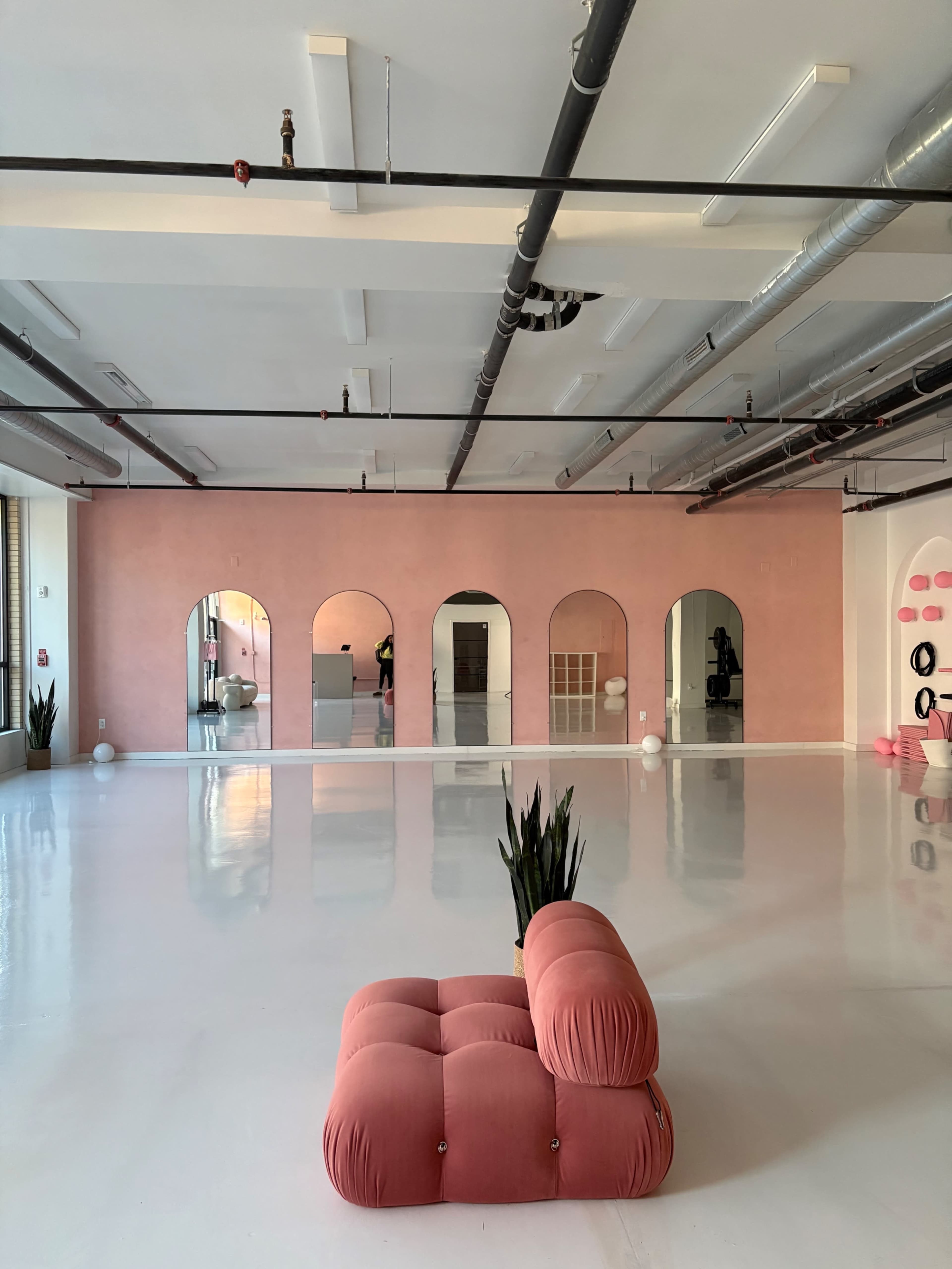 The image shows a spacious interior with a polished white floor, pink walls, large arched mirrors, and a pink tufted couch positioned in the center.