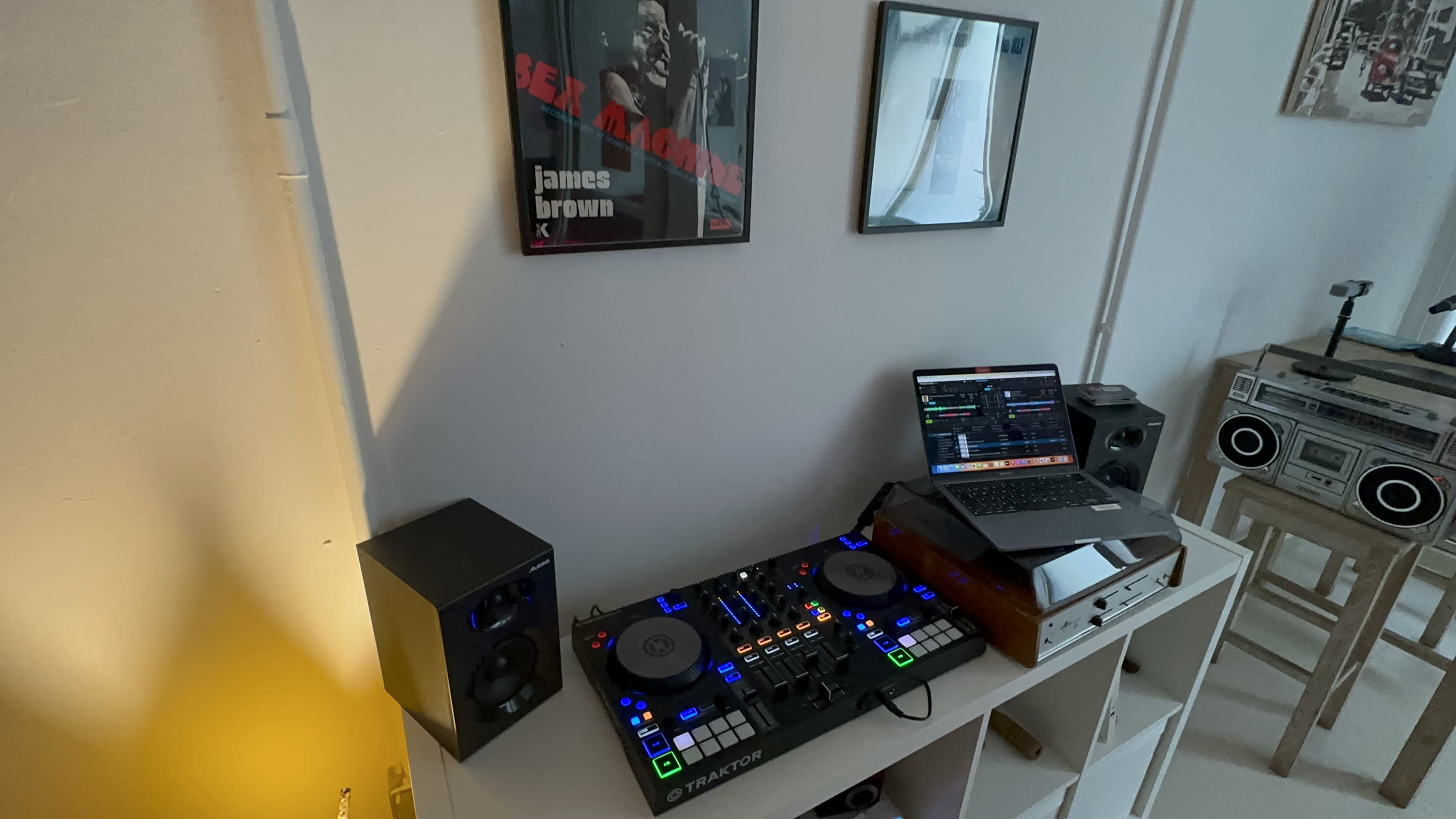 A digital music setup featuring a DJ controller, a laptop displaying software, speakers, and framed posters on the wall.