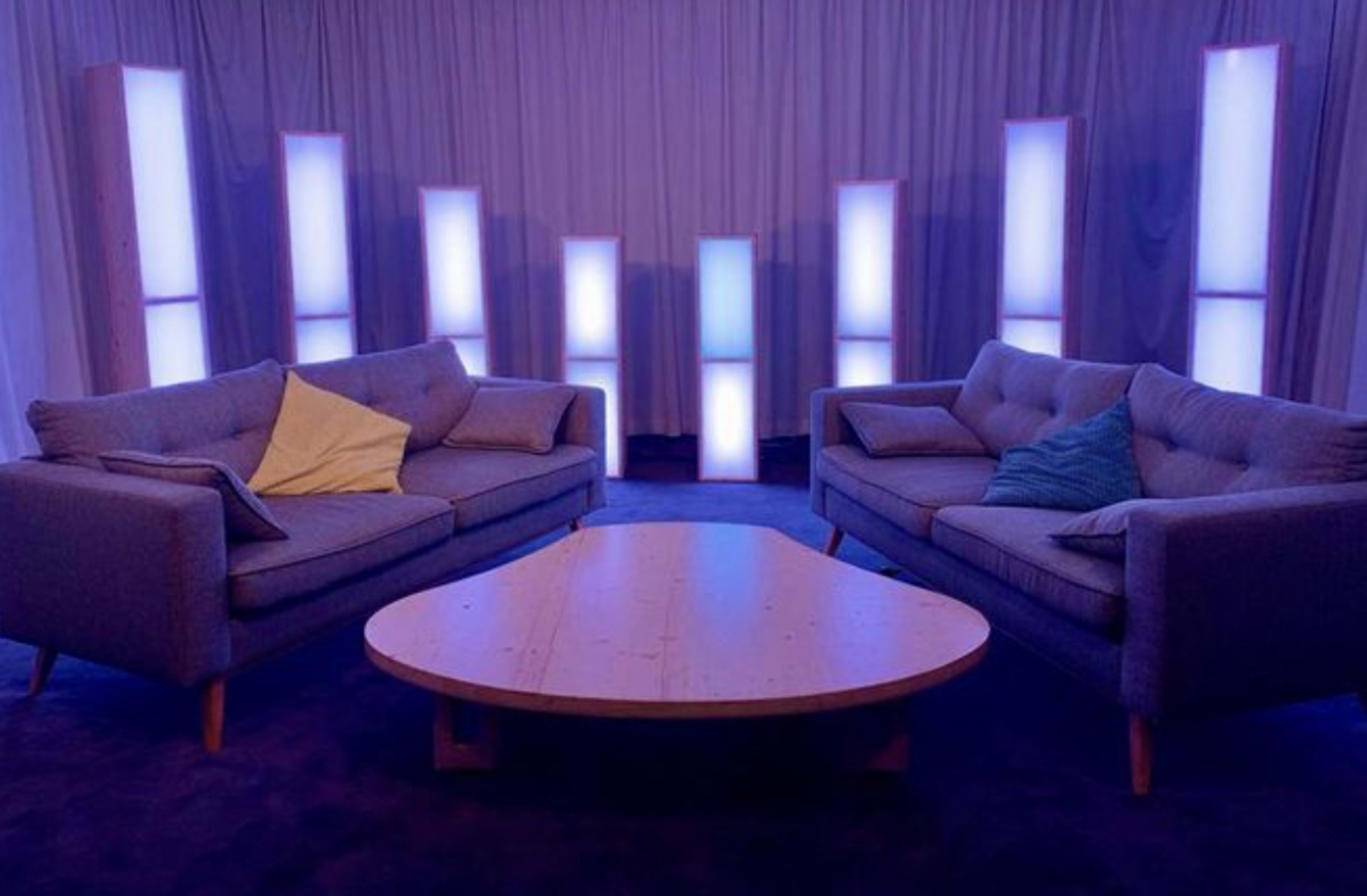 Two gray sofas facing each other across a wooden coffee table, surrounded by illuminated vertical panels in a softly lit room.