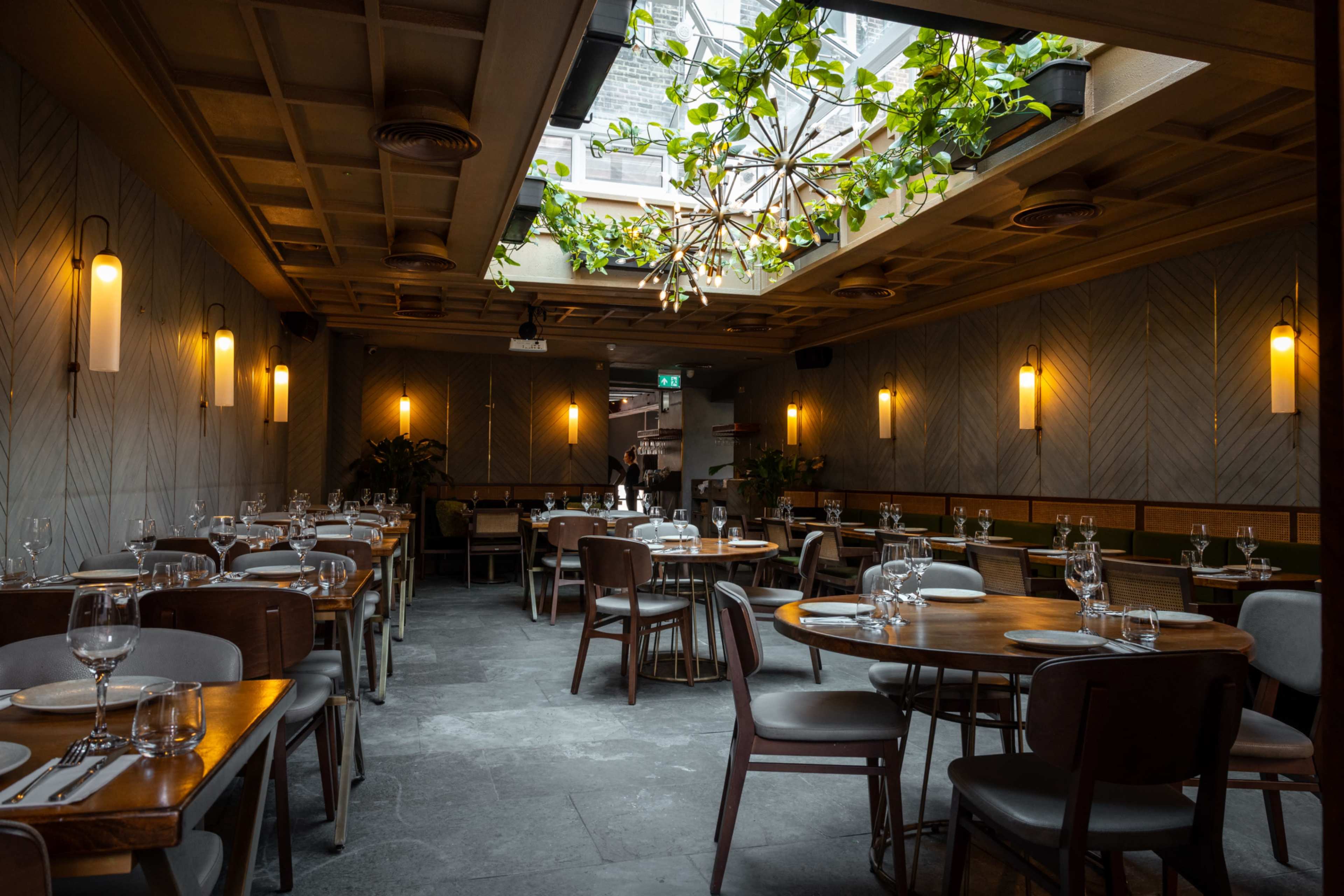 The image shows a modern restaurant interior with wooden tables, upholstered chairs, and a skylight allowing natural light to enter.