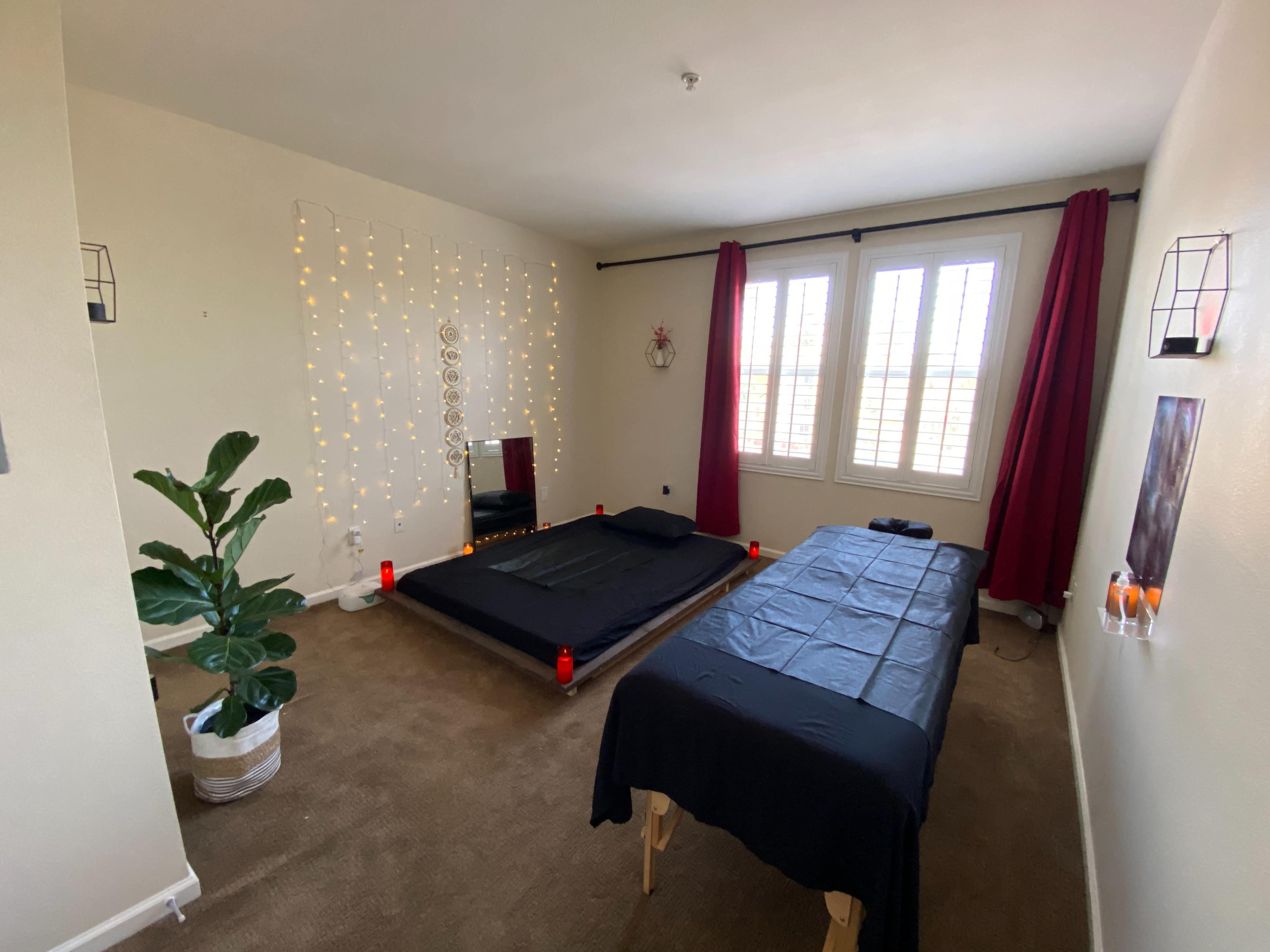 The room features a low black bed surrounded by candles, a table covered with a black cloth, and large windows with red curtains.