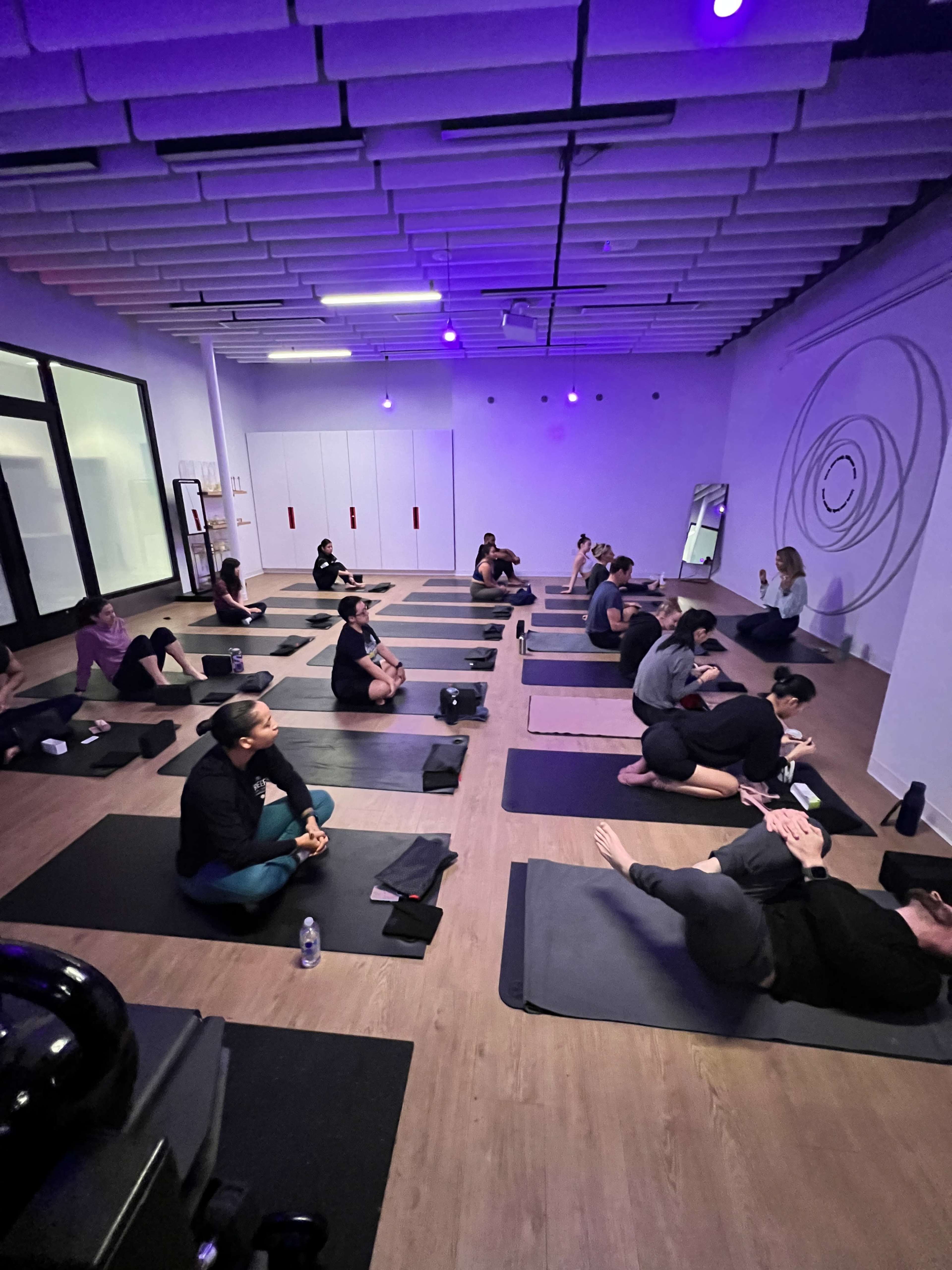 A group of people is seated on yoga mats in a studio with purple lighting.