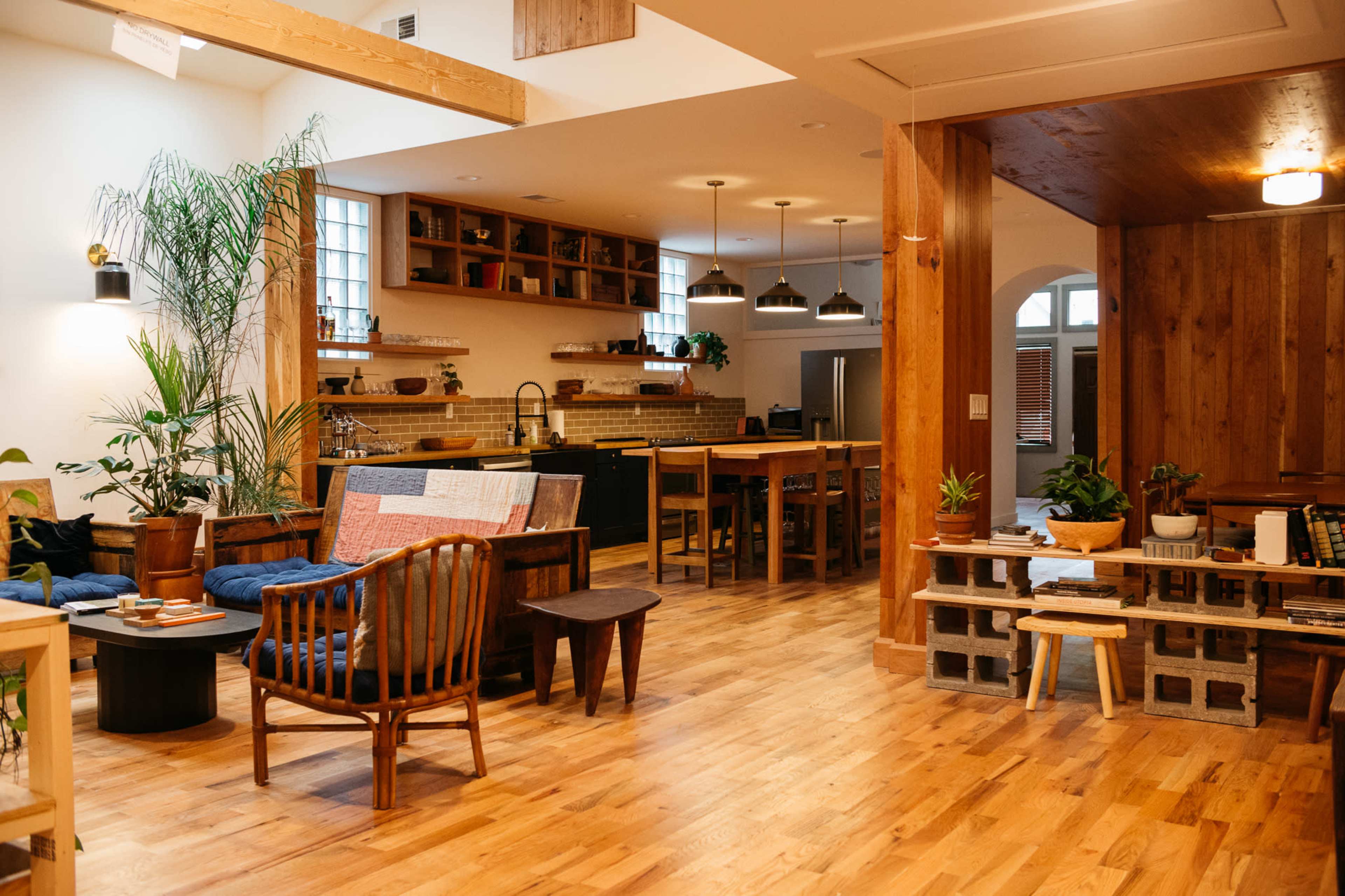 A modern, open-concept living space featuring wooden floors, a kitchen area with shelves, dining tables, and various plants throughout the room.