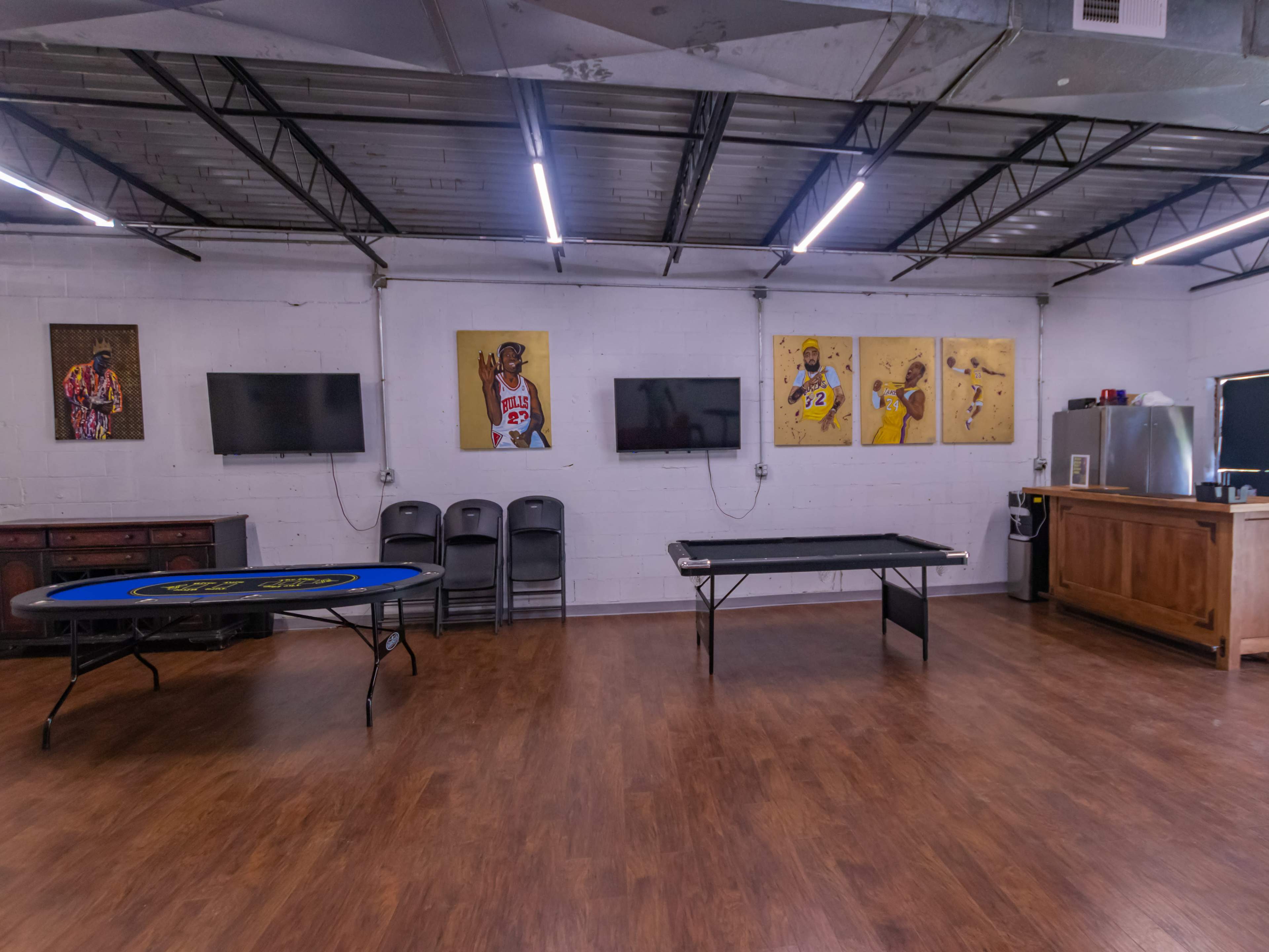 A spacious recreational room with two tables, several chairs, television screens, and framed basketball artwork on the walls.