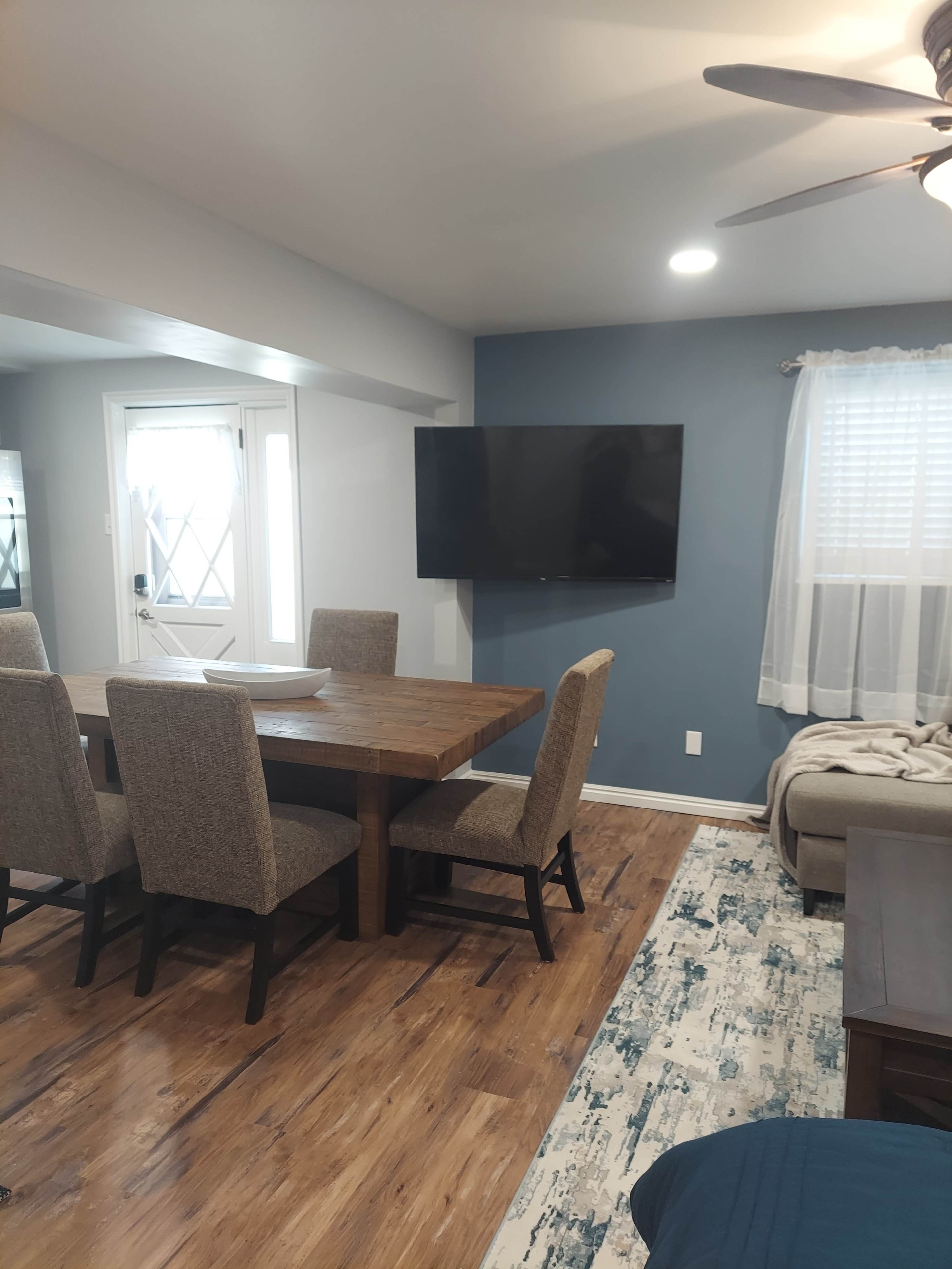 A dining area with a wooden table and six upholstered chairs, adjacent to a living space featuring a sofa, television mounted on a blue wall, and a window with sheer curtains.
