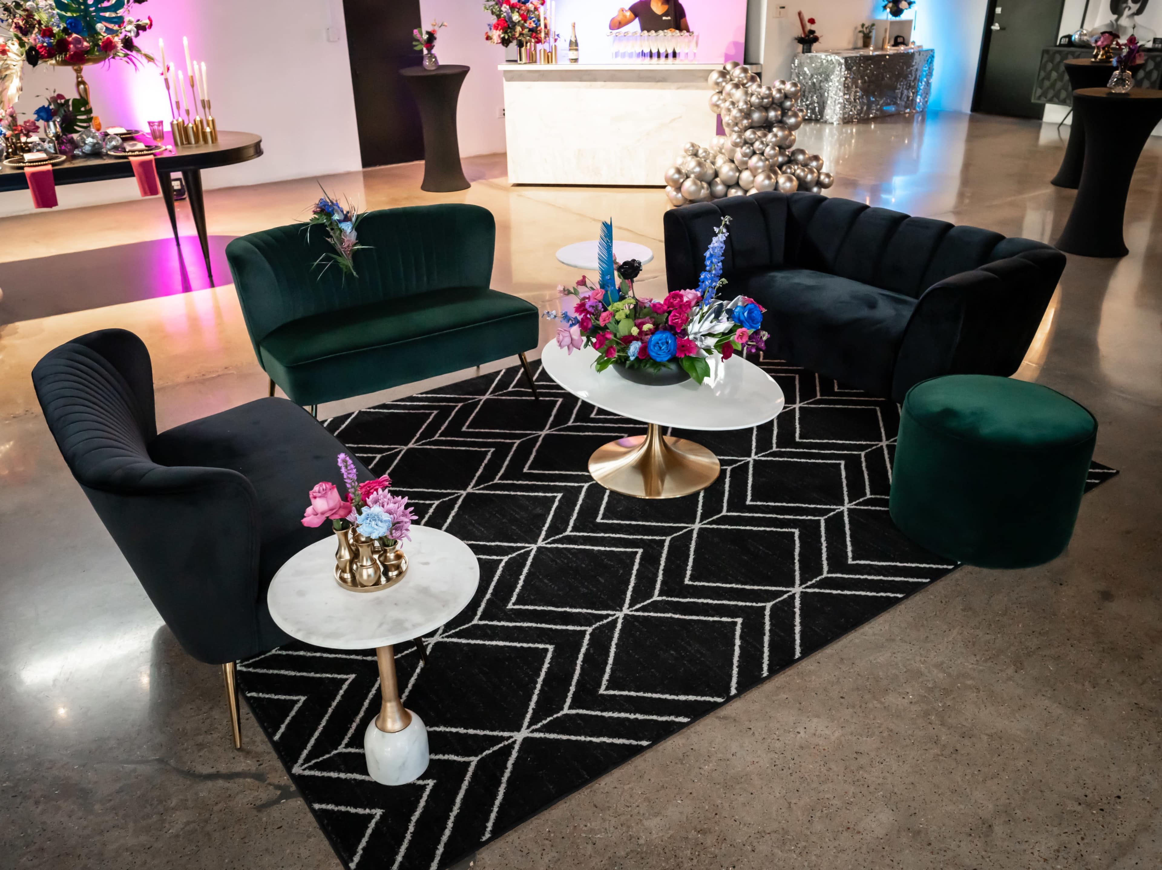 A modern lounge area featuring two velvet sofas, a round stool, and marble-top tables, all arranged on a geometric-patterned rug with floral centerpieces.