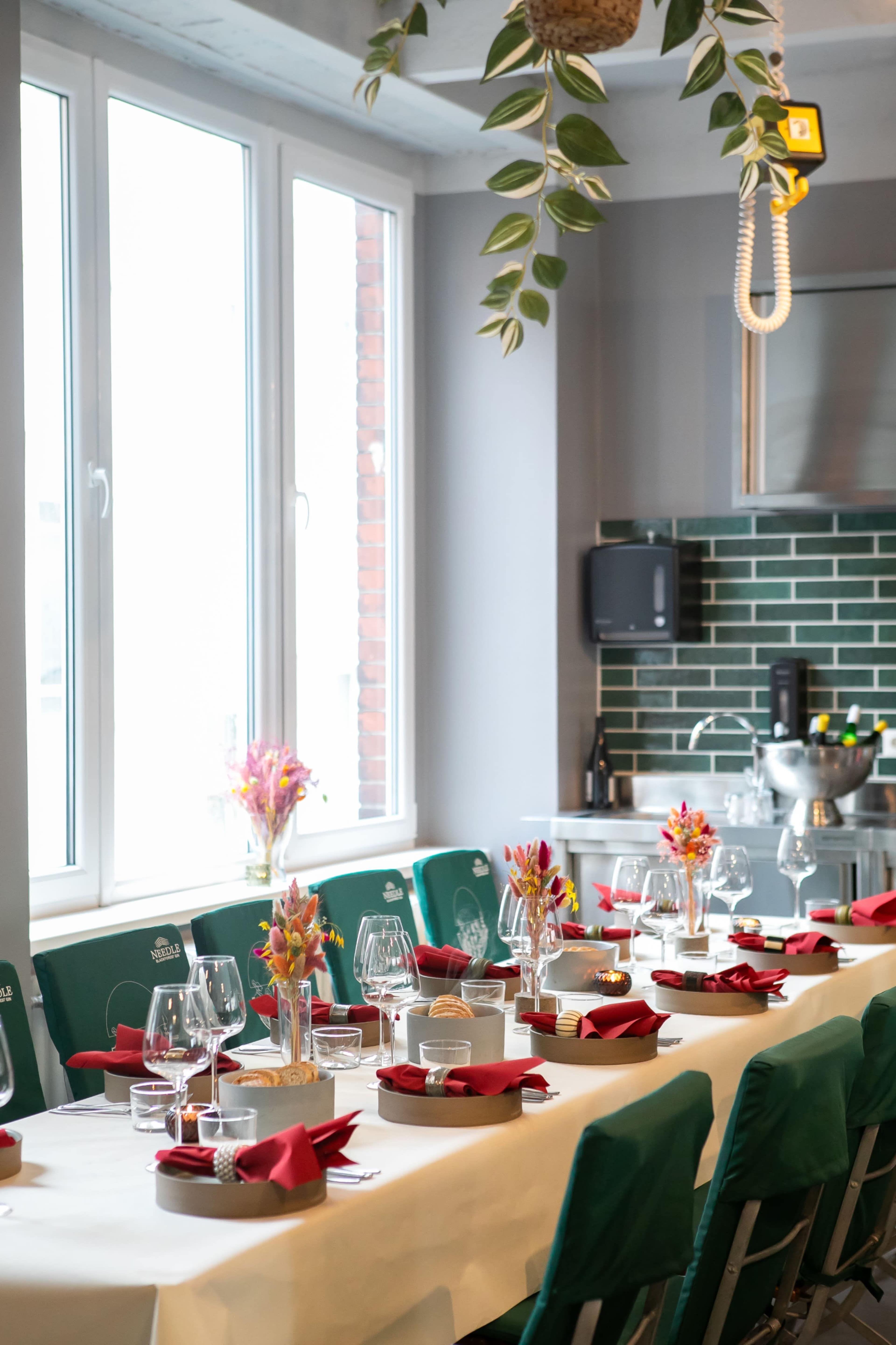 A long dining table is set with glasses, plates, and red napkins, positioned near large windows that let in natural light.