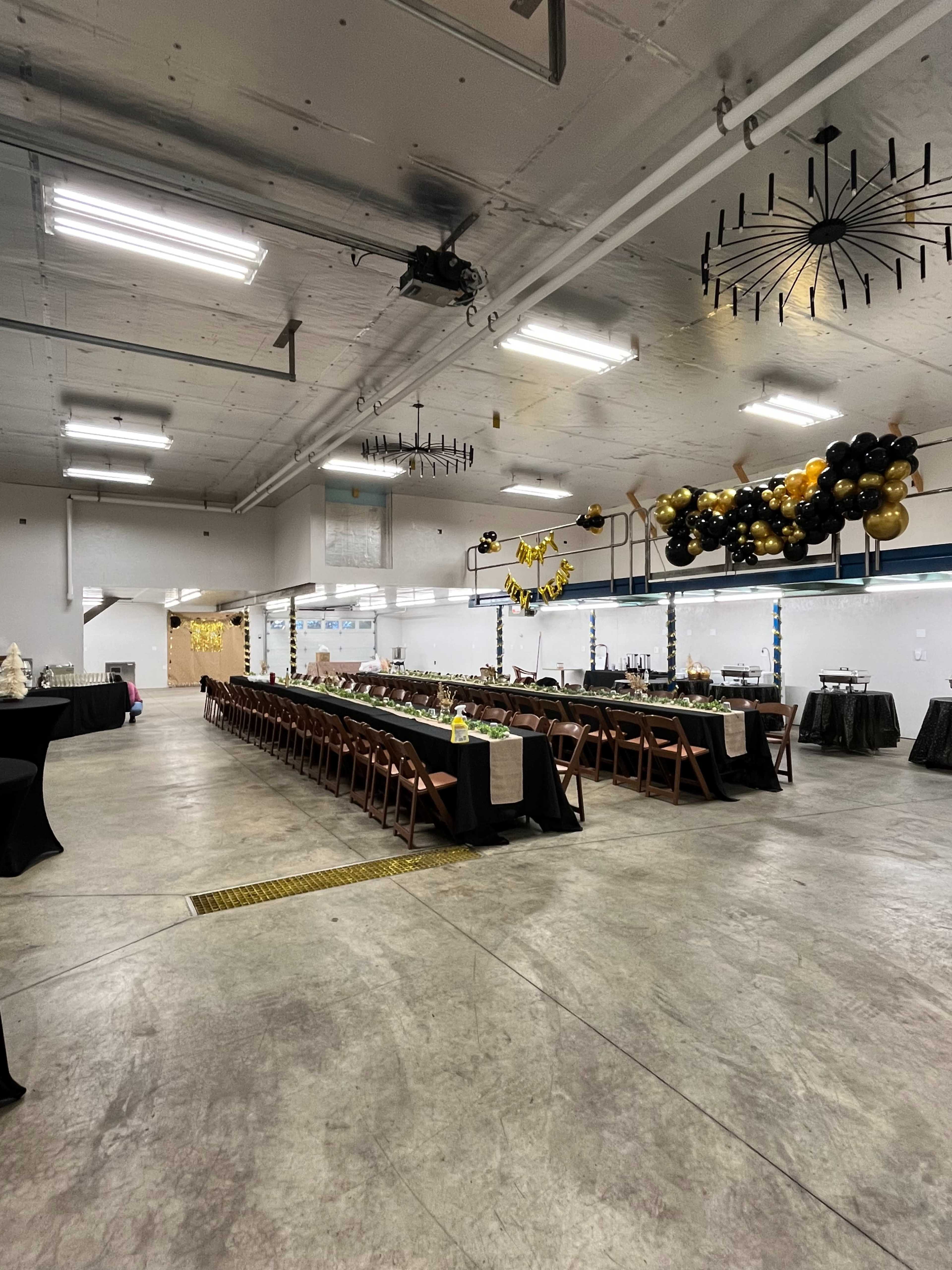 A spacious indoor venue features long tables arranged for a banquet, decorated with black and gold balloons and additional tables set for serving.