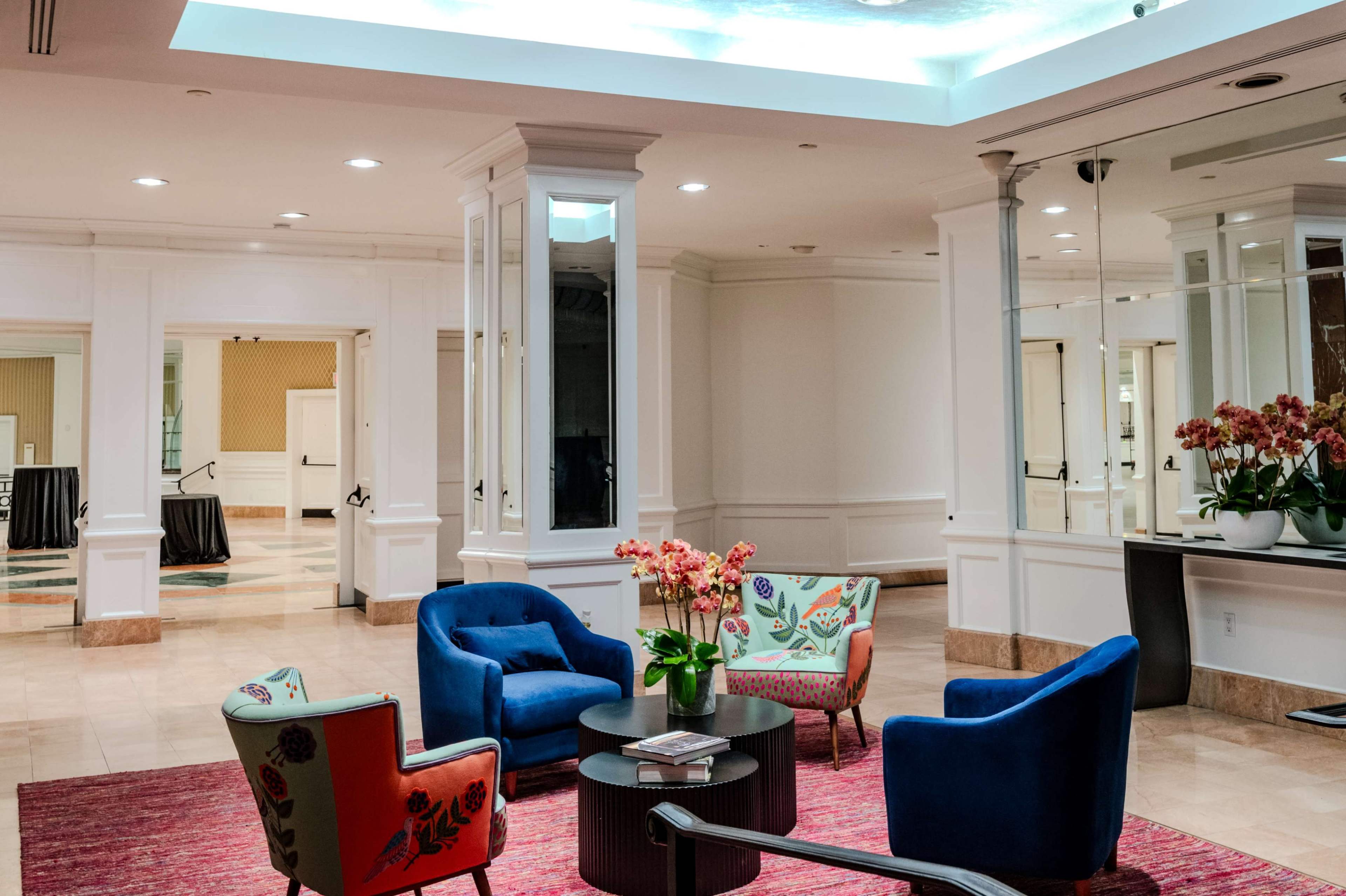 The image shows a modern lobby area featuring blue upholstered chairs, patterned armchairs, a round coffee table, and a floral arrangement.