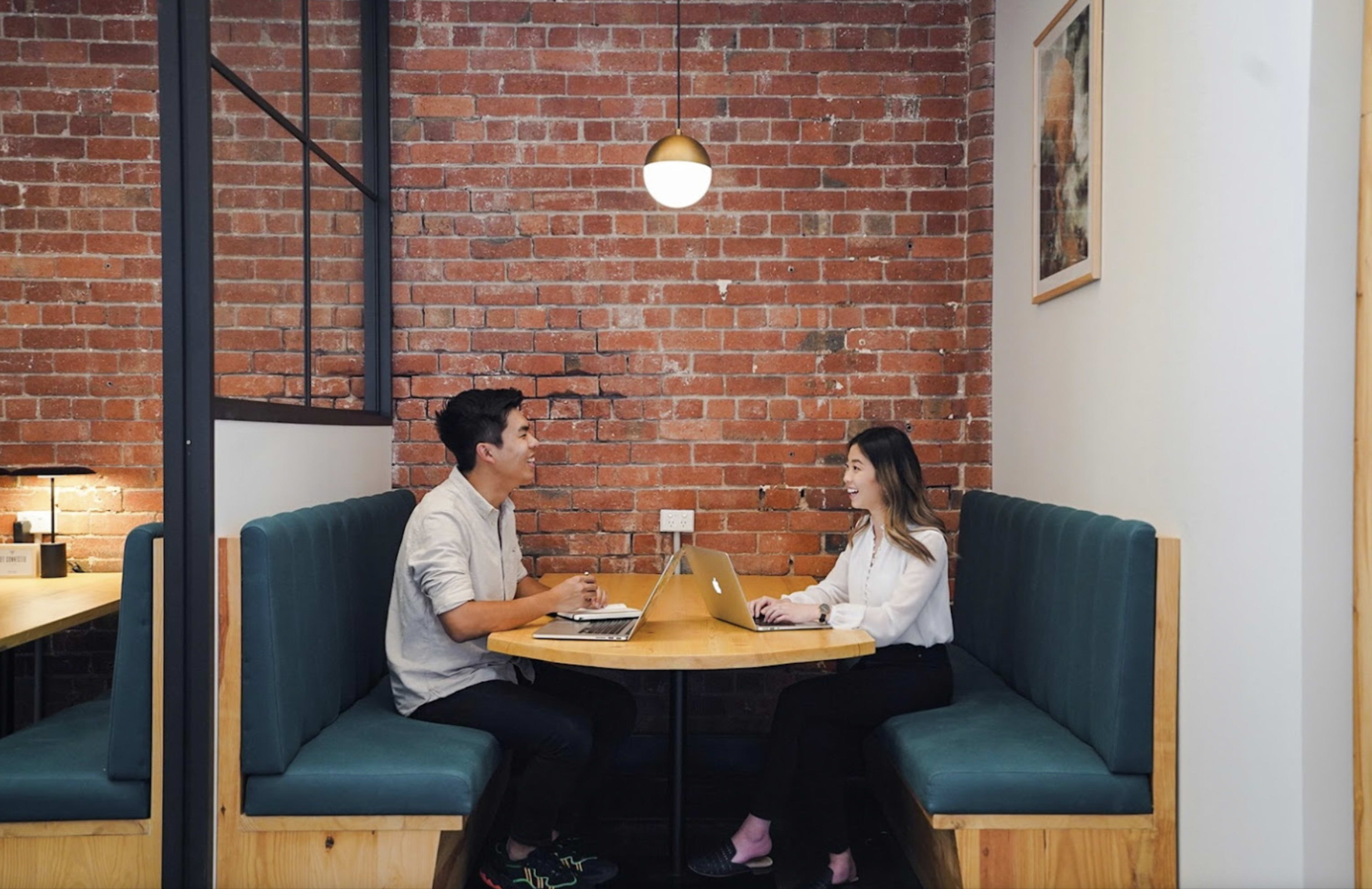 Two people are seated at a round table in a cafe, engaged in conversation while using a laptop.