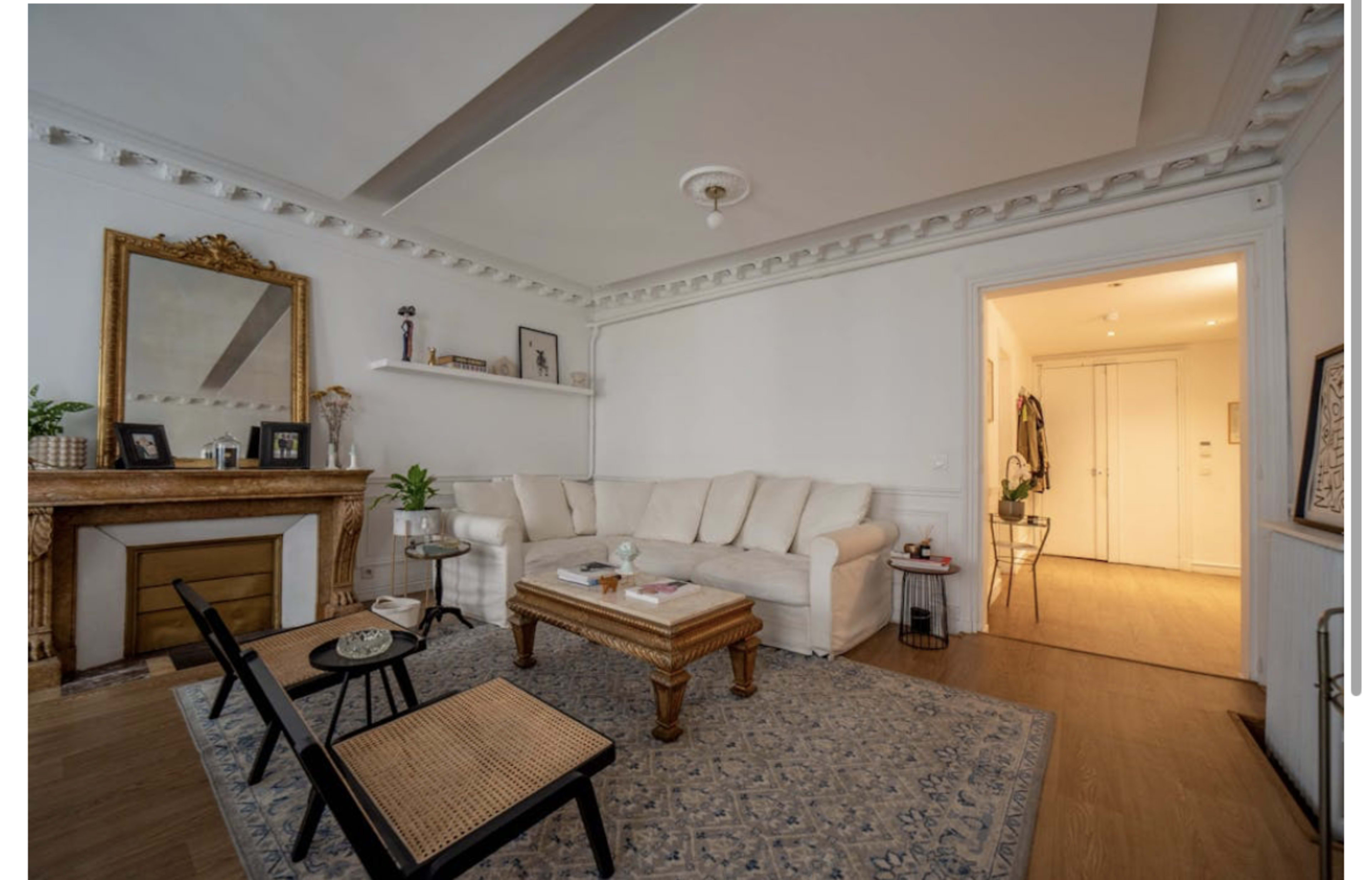 The image shows a bright living room with a beige sofa, a coffee table, and a decorative fireplace, which is connected to a hallway leading to other rooms.