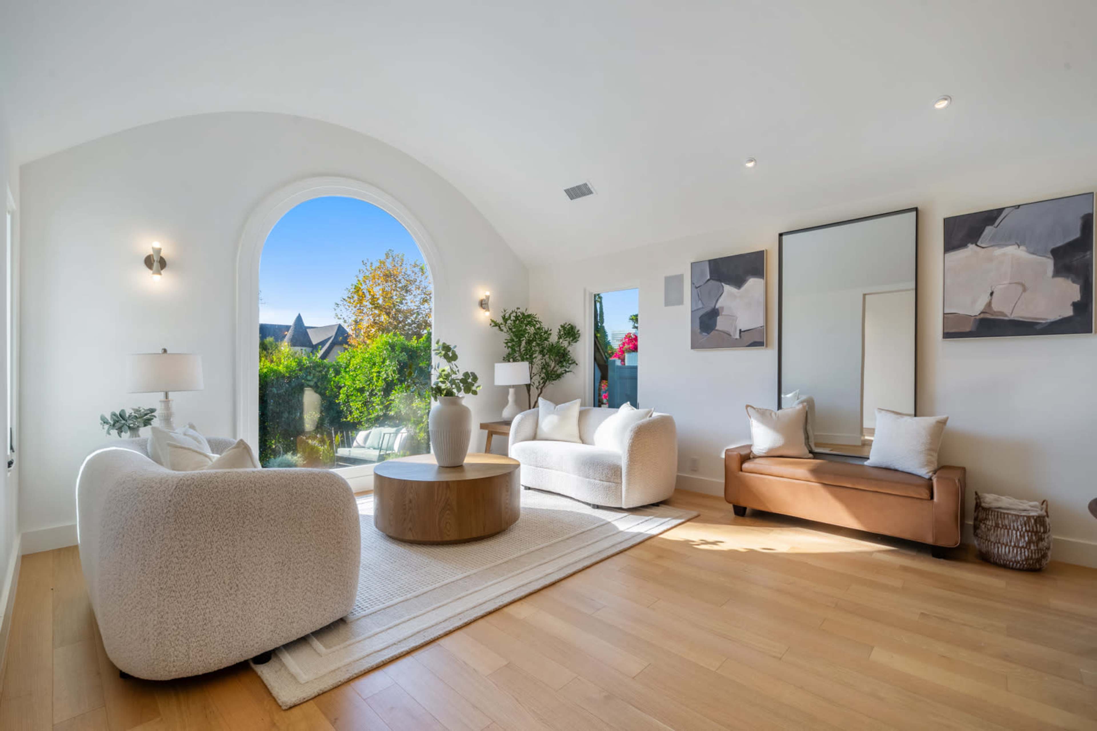 The image shows a bright living room featuring curved white sofas, a round wooden coffee table, and a large arched window with a view of lush greenery.